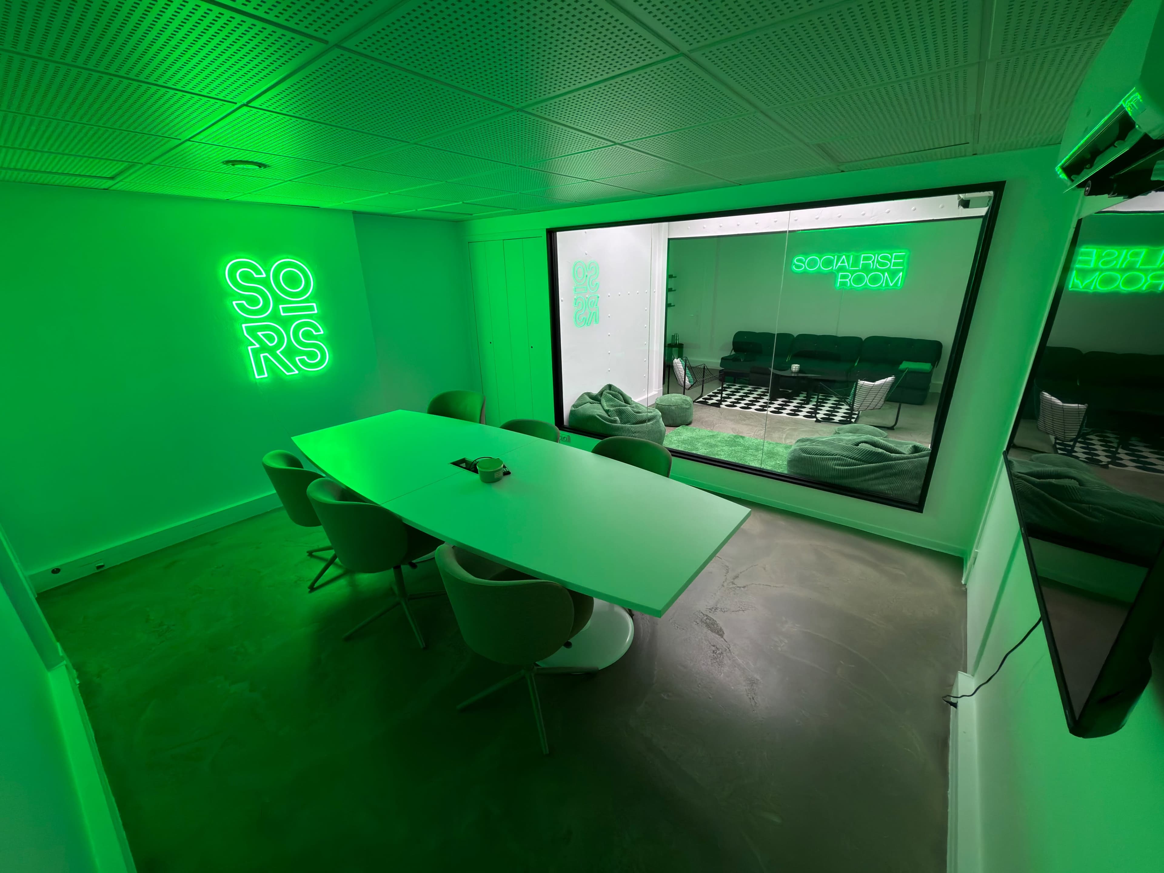 The image shows a modern meeting room with a large white table, chairs, and neon green signage on the walls.