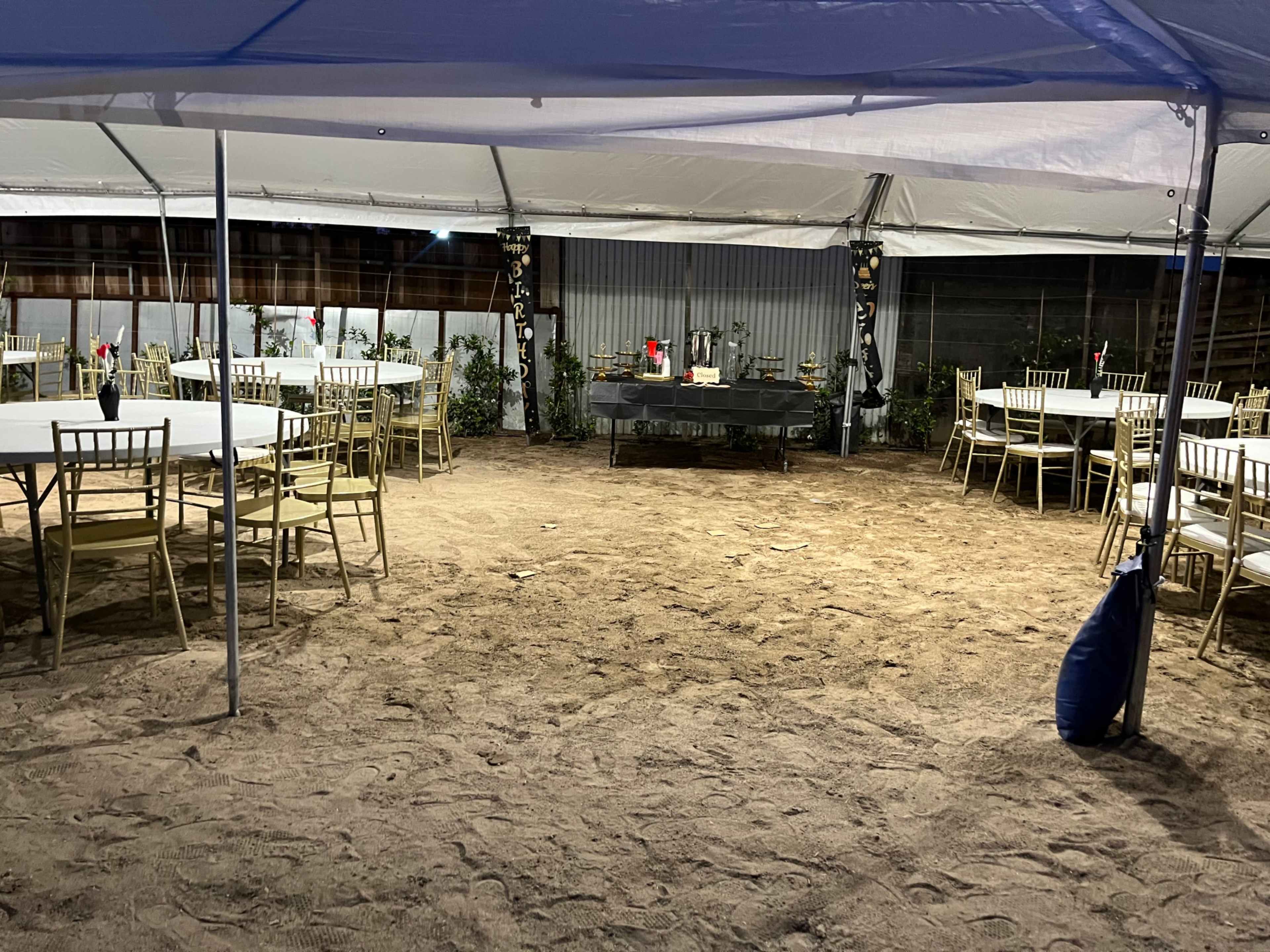 A large tented area features circular tables and chairs arranged on sandy ground, with a decorative table at the center surrounded by plants and flowers.