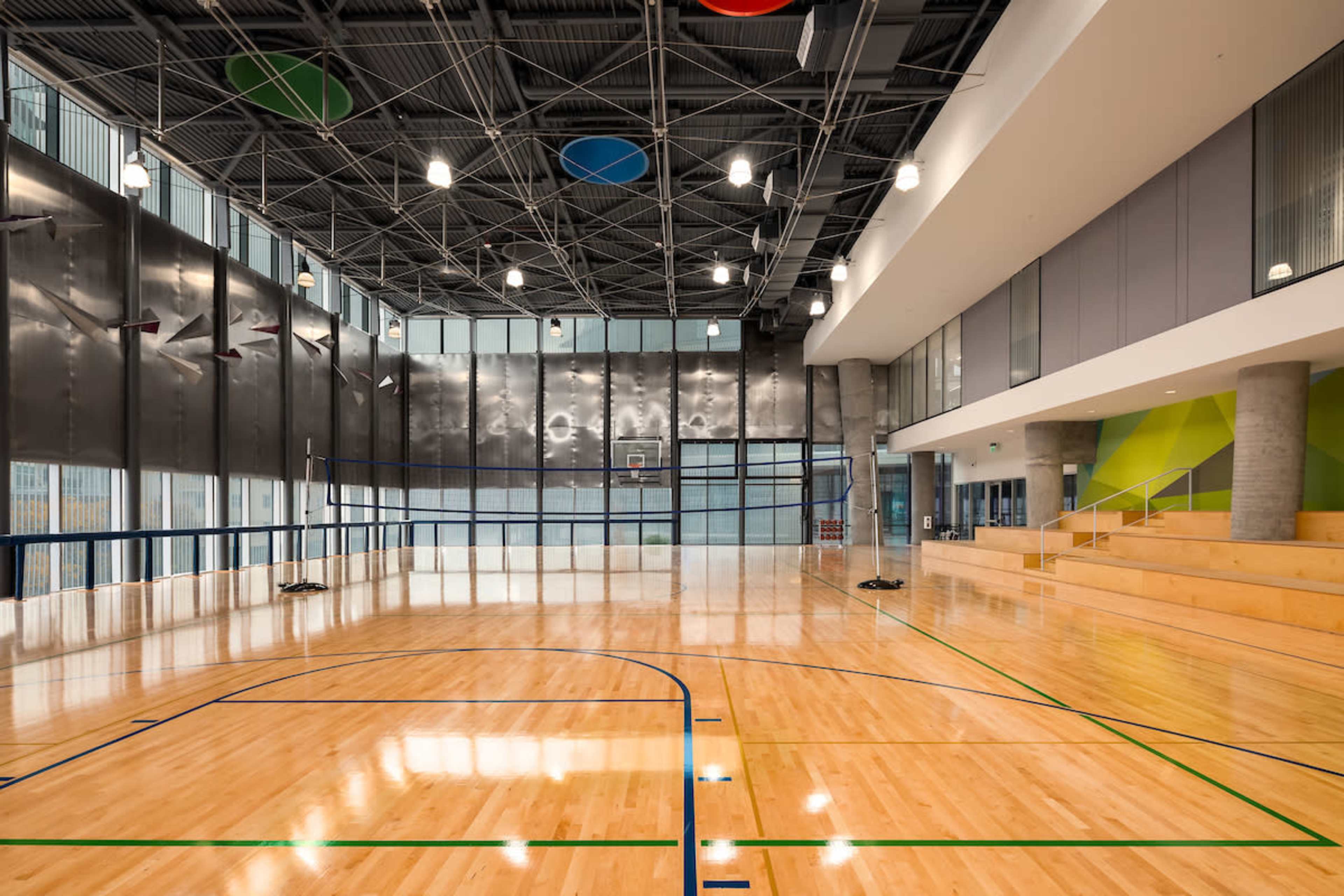 A spacious gymnasium with polished wooden floors, large windows, and areas for basketball and seating.