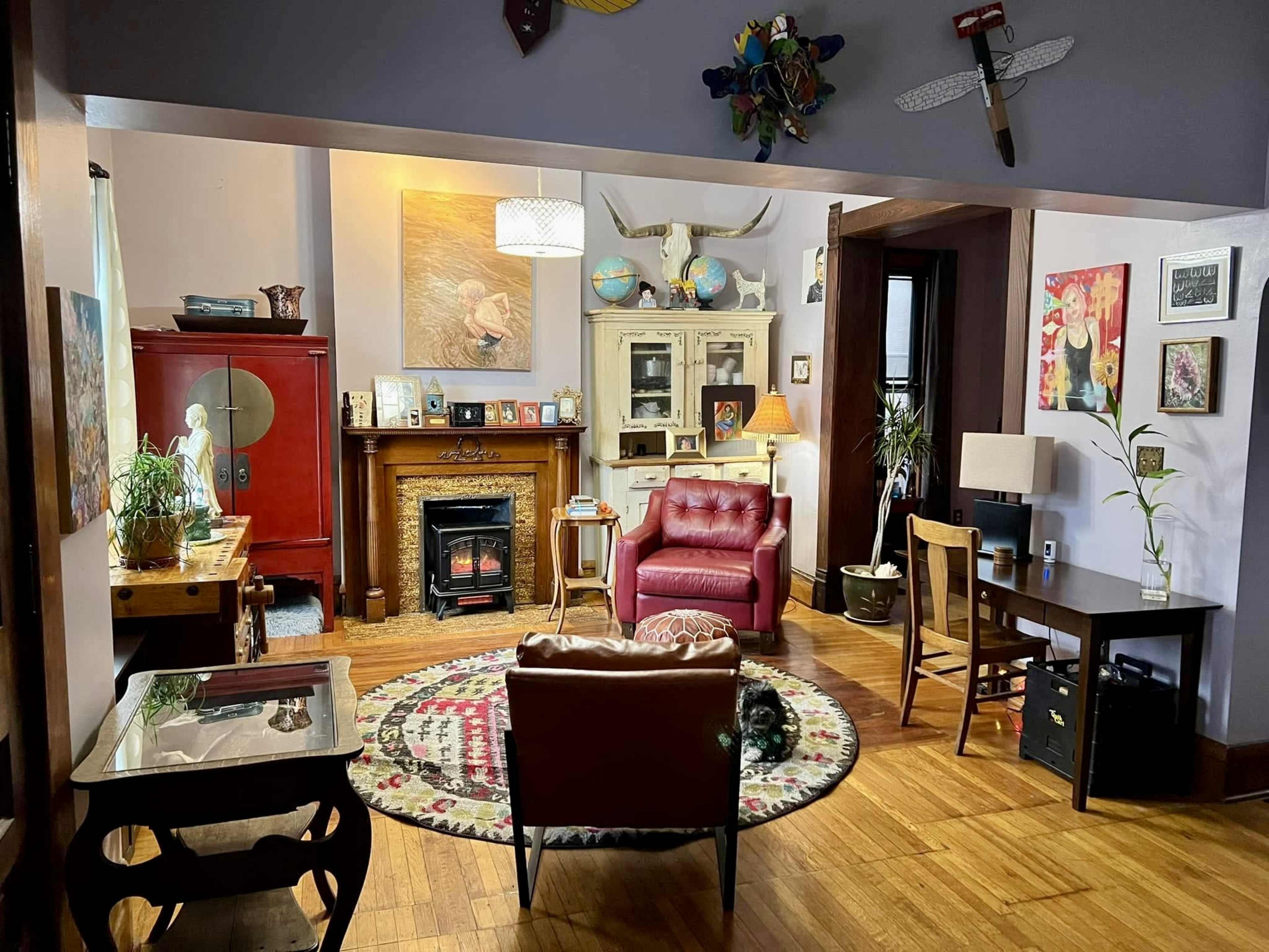 A cozy living room features a red leather armchair, a patterned area rug, a small desk, and a fireplace surrounded by eclectic decor.
