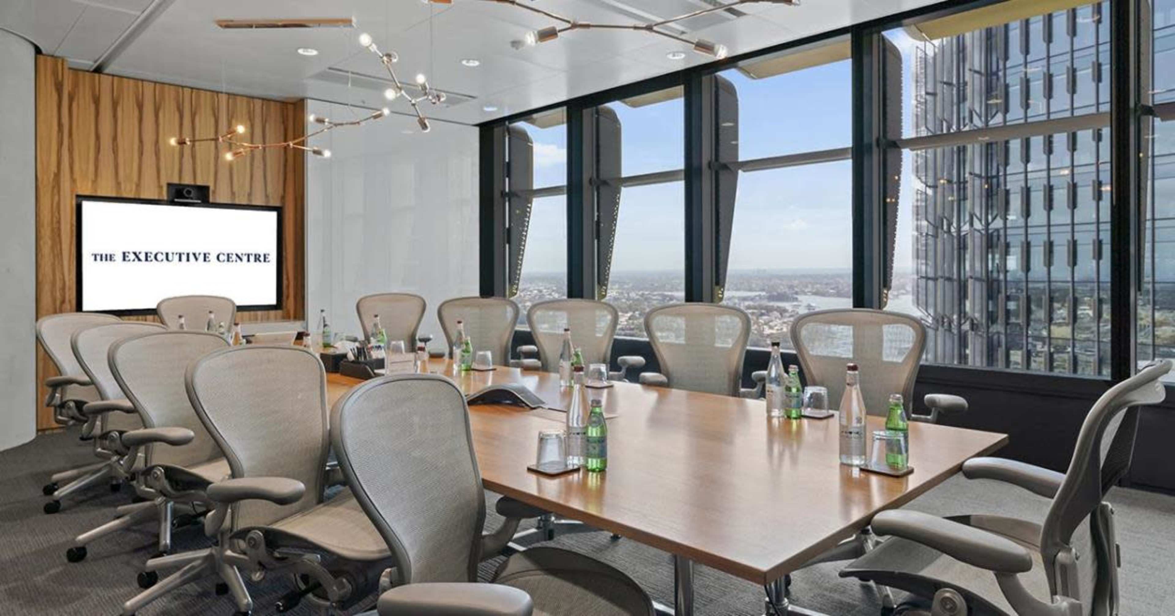 A modern executive conference room features a large wooden table surrounded by ergonomic chairs, with a screen displaying "The Executive Centre" and large windows offering a city view.