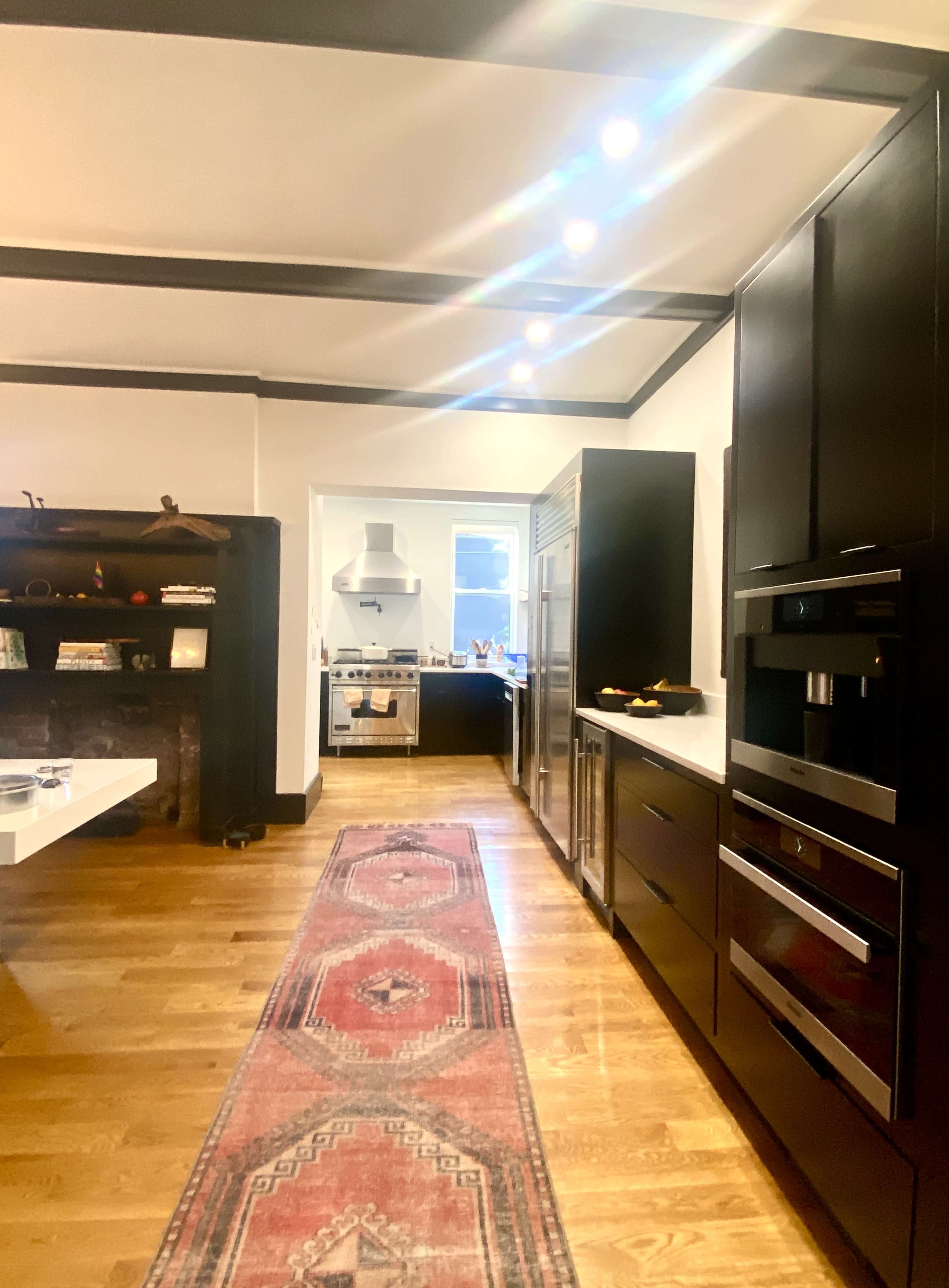 A modern kitchen features dark cabinetry, stainless steel appliances, and a central island, with a patterned runner rug extending down the wooden floor.