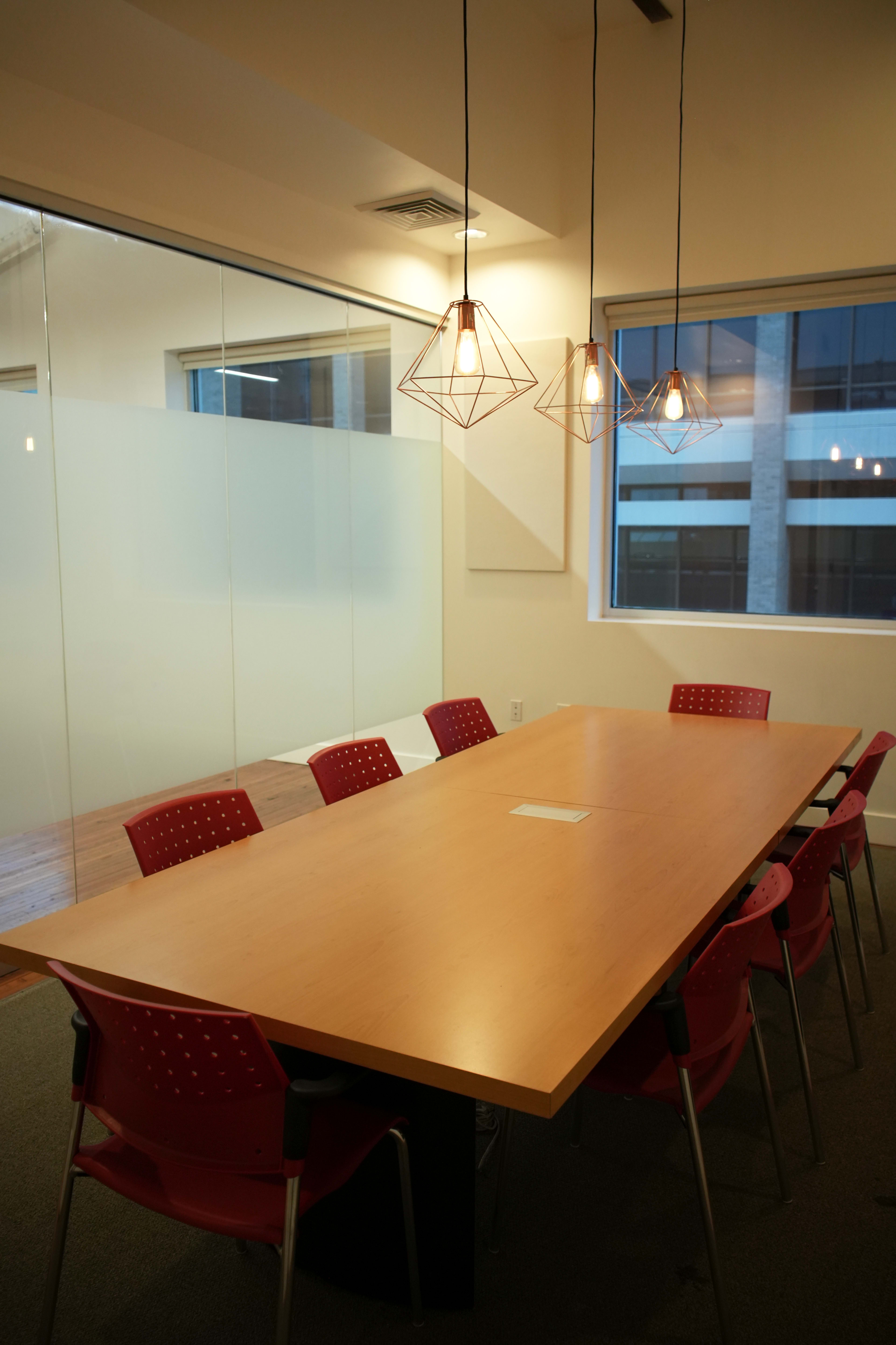 A long wooden conference table surrounded by red chairs is illuminated by geometric pendant lights in a modern meeting room.