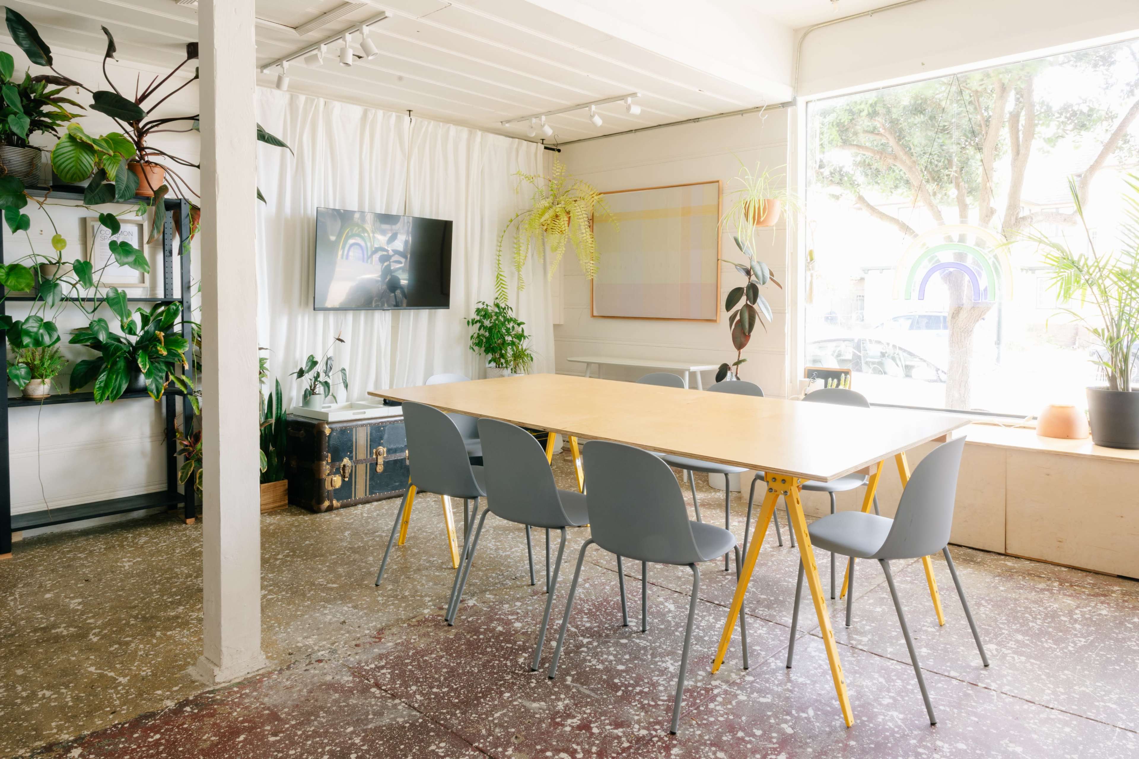 A spacious meeting room features a long wooden table surrounded by gray chairs, with plants and a television set in the background.