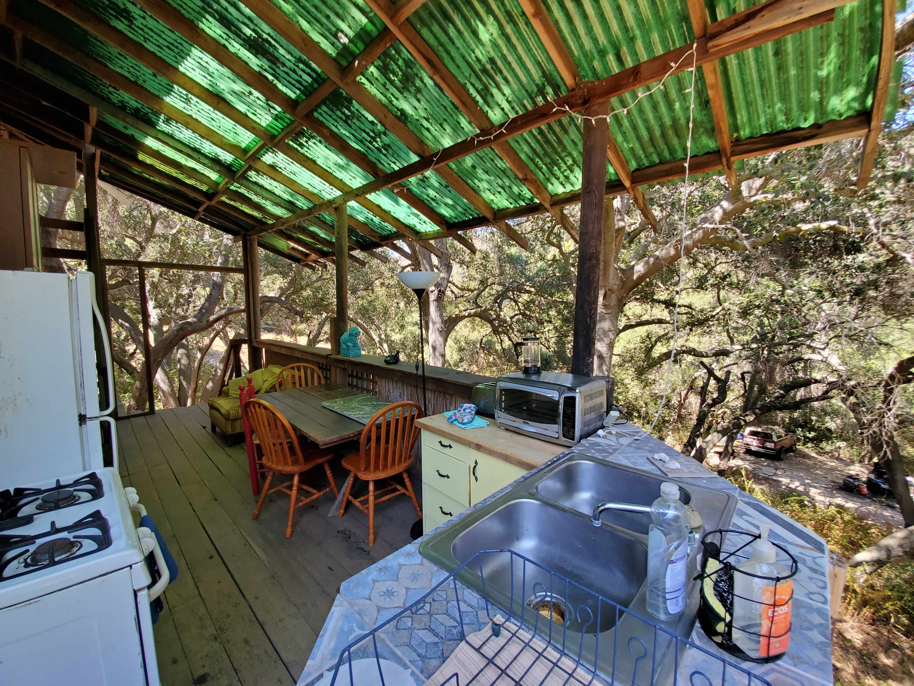 A rustic kitchen area with a wooden table and chairs, surrounded by trees, featuring a stove, sink, and a microwave under a green corrugated roof.