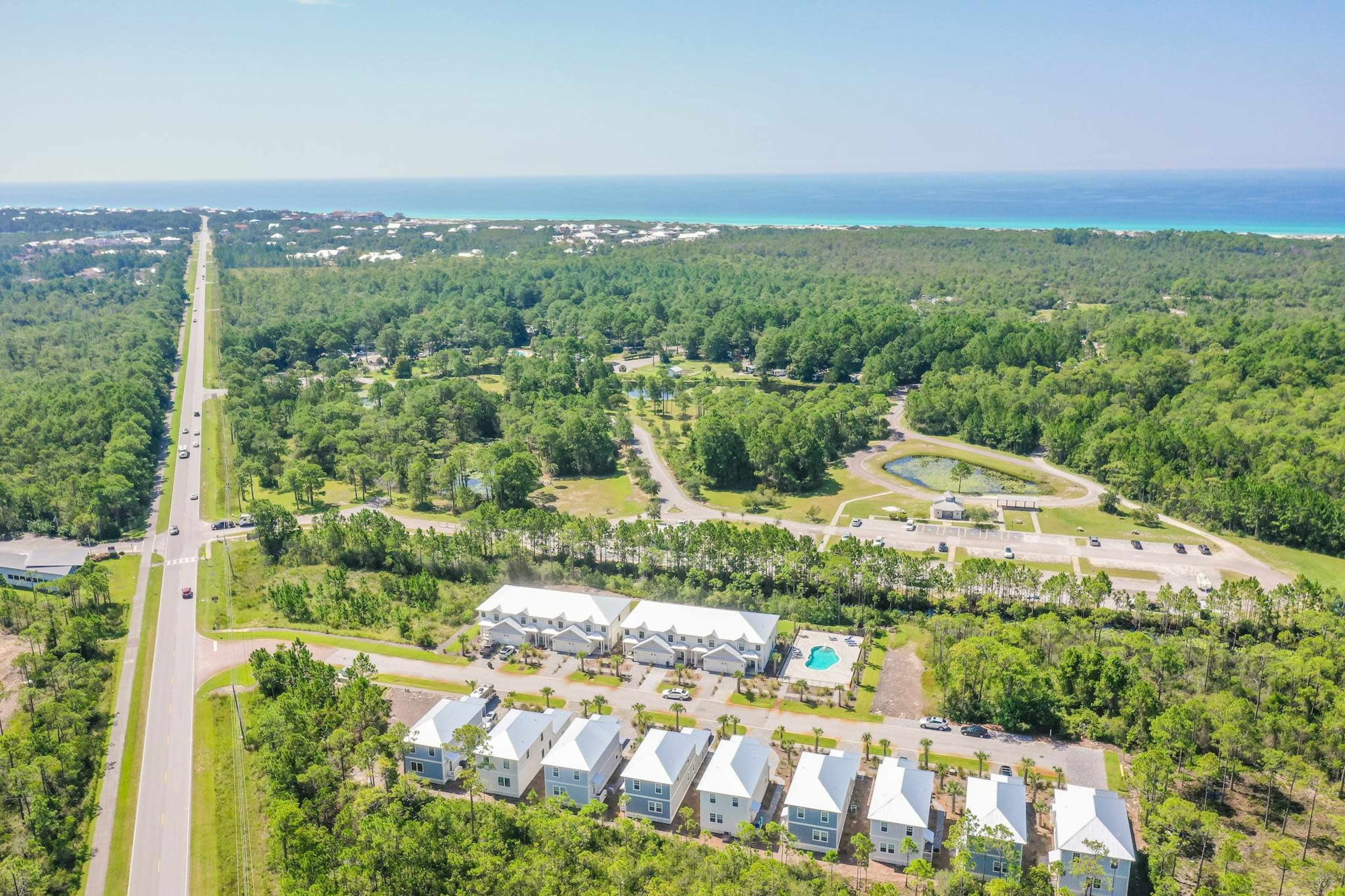 A view of a residential area with several clustered houses near a highway, surrounded by green trees and close to a coastline in the distance.