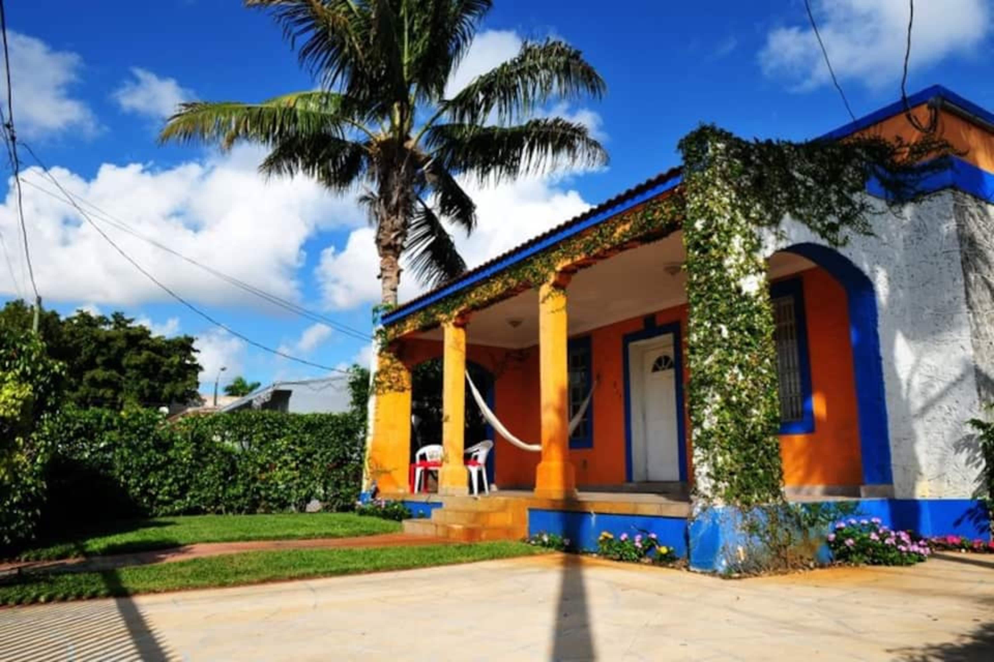 A colorful house with orange and blue accents, featuring columns and a porch with two white chairs, surrounded by greenery and a palm tree.