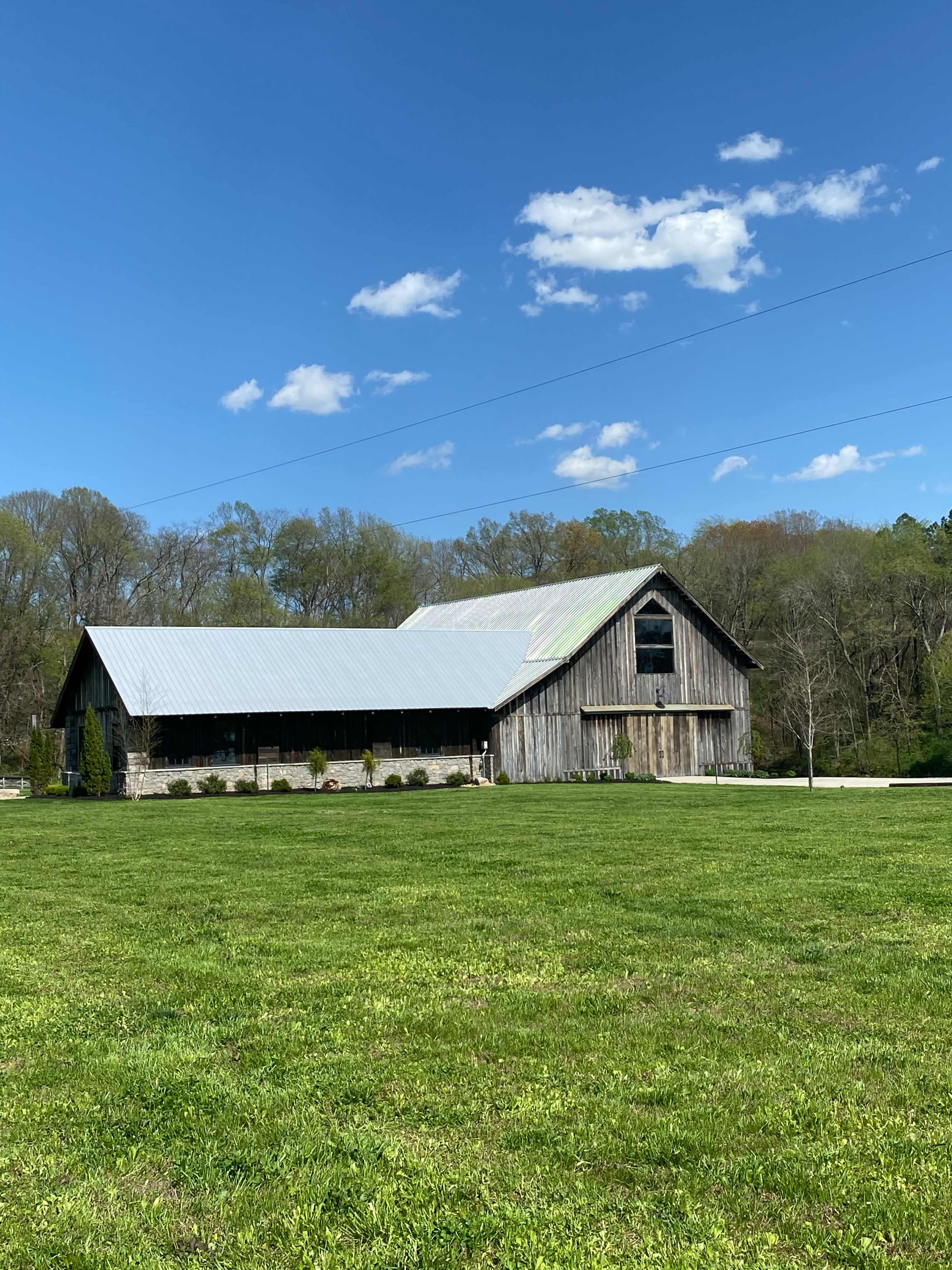 A wooden barn with a metal roof stands beside a green lawn under a blue sky with scattered clouds.