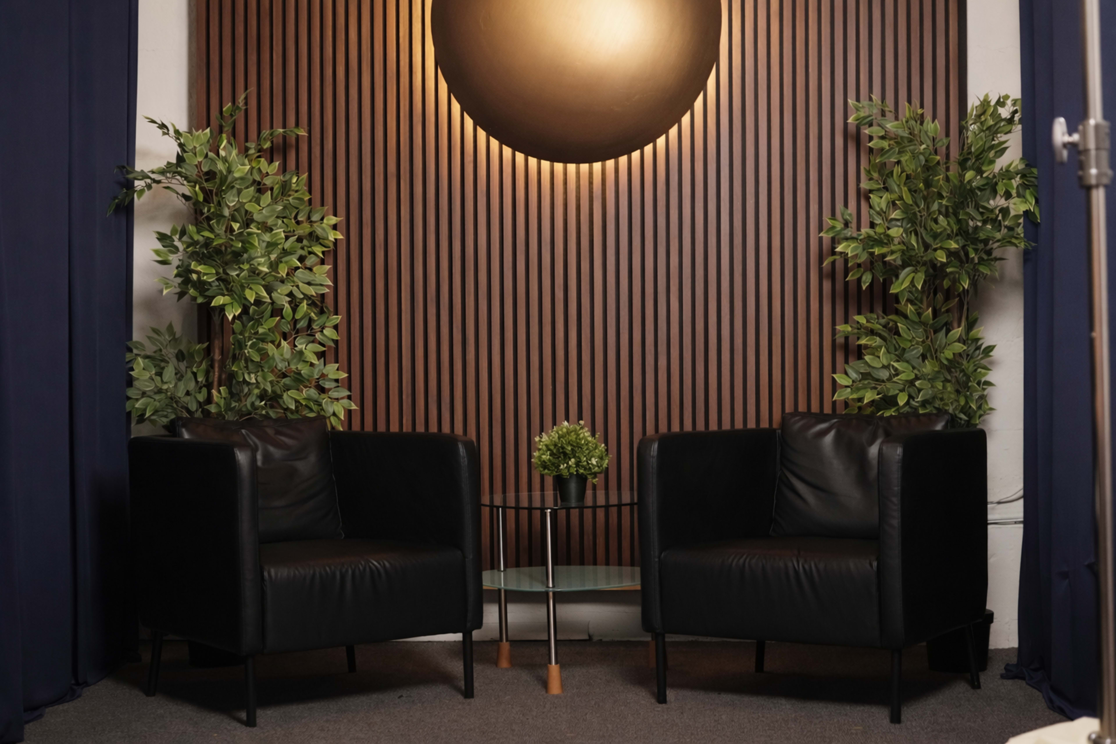 Two black leather chairs are positioned on either side of a small glass table, surrounded by leafy plants against a wooden panel wall with a circular light fixture above.