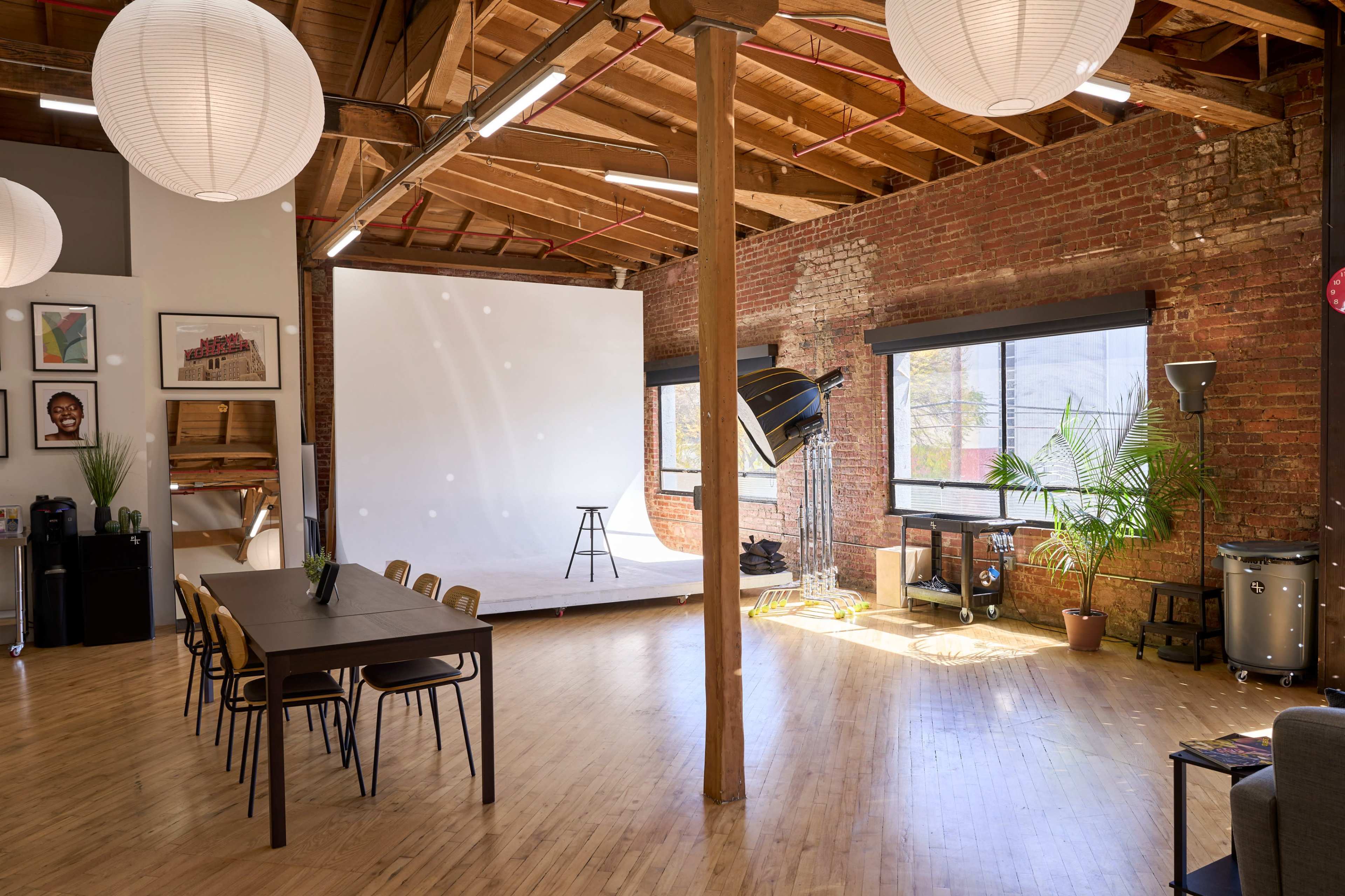 The image shows a spacious studio with exposed brick walls, wooden beams, and a large white backdrop, featuring a dining table with chairs and a lighting setup in the corner.