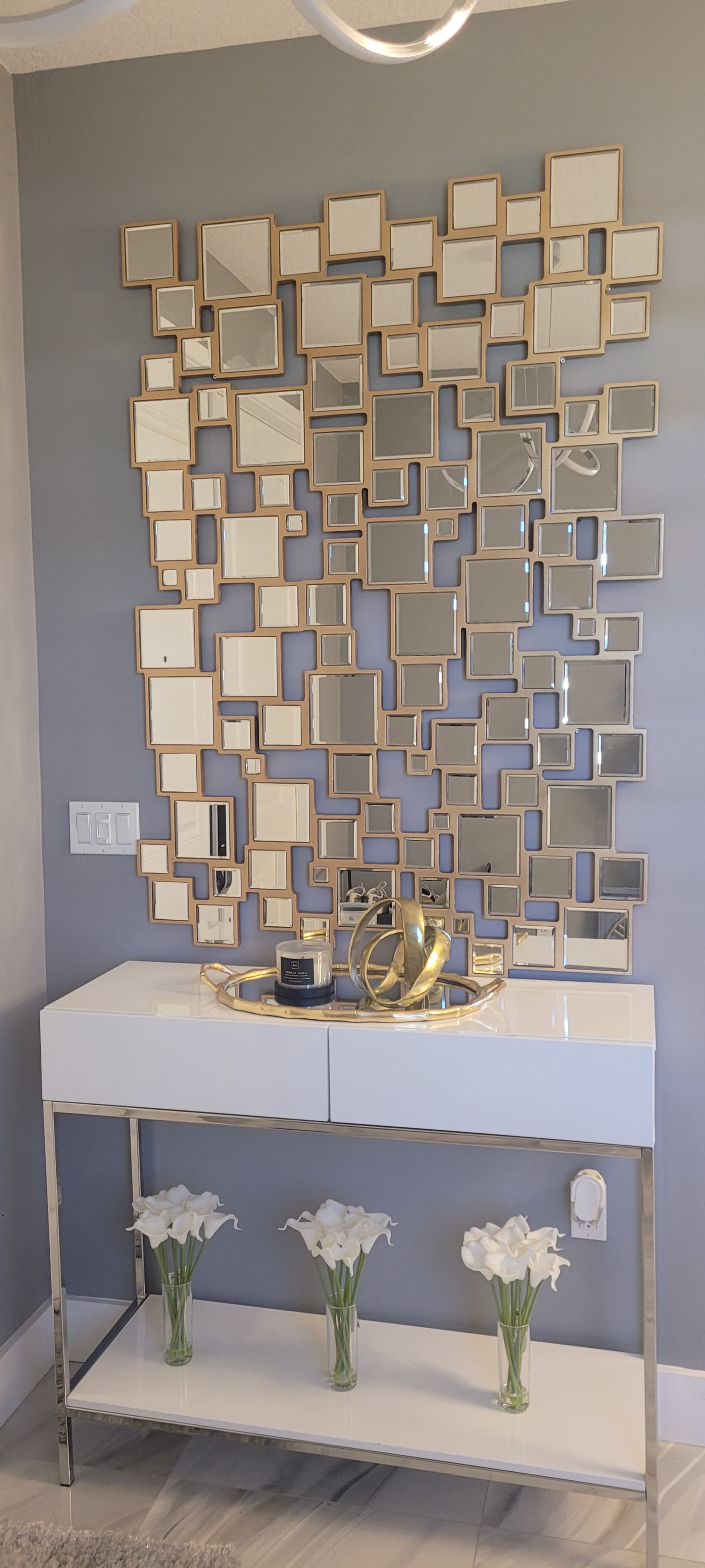A modern entryway with a wall-mounted arrangement of small, framed mirrors above a sleek white console table adorned with a decorative tray and three vases of white flowers.
