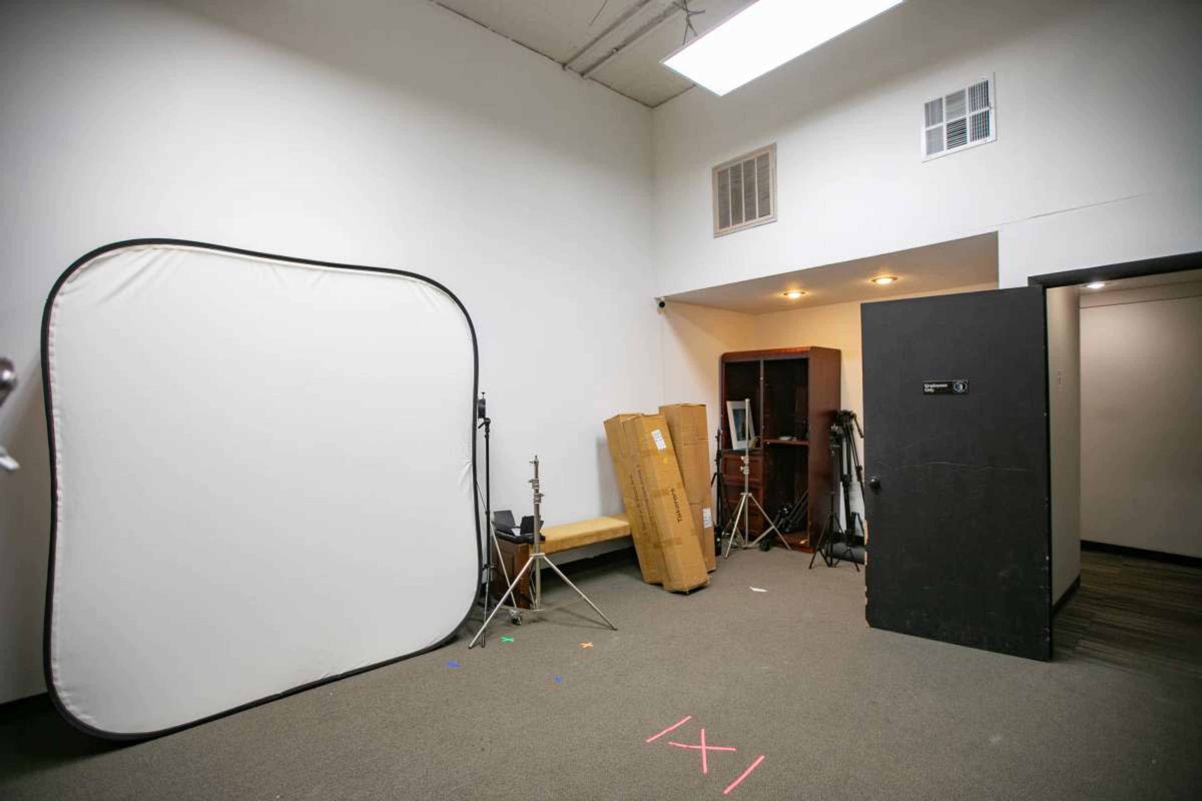 The image shows a vacant studio space featuring a large white backdrop, a few equipment stands, a cardboard box, and a black partition against the wall.