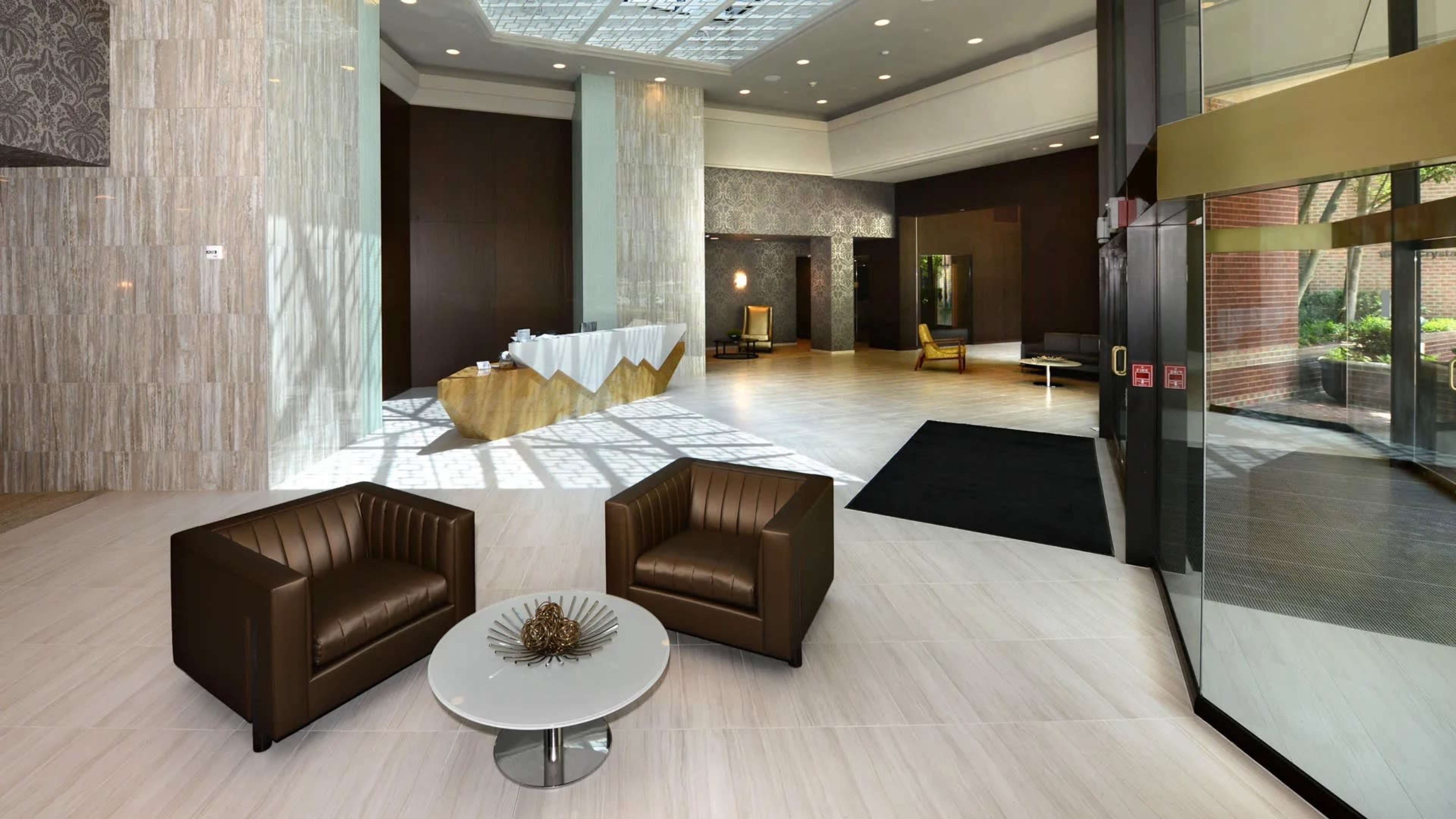 A spacious hotel lobby with two brown leather chairs, a round coffee table, and a reception area in the background.