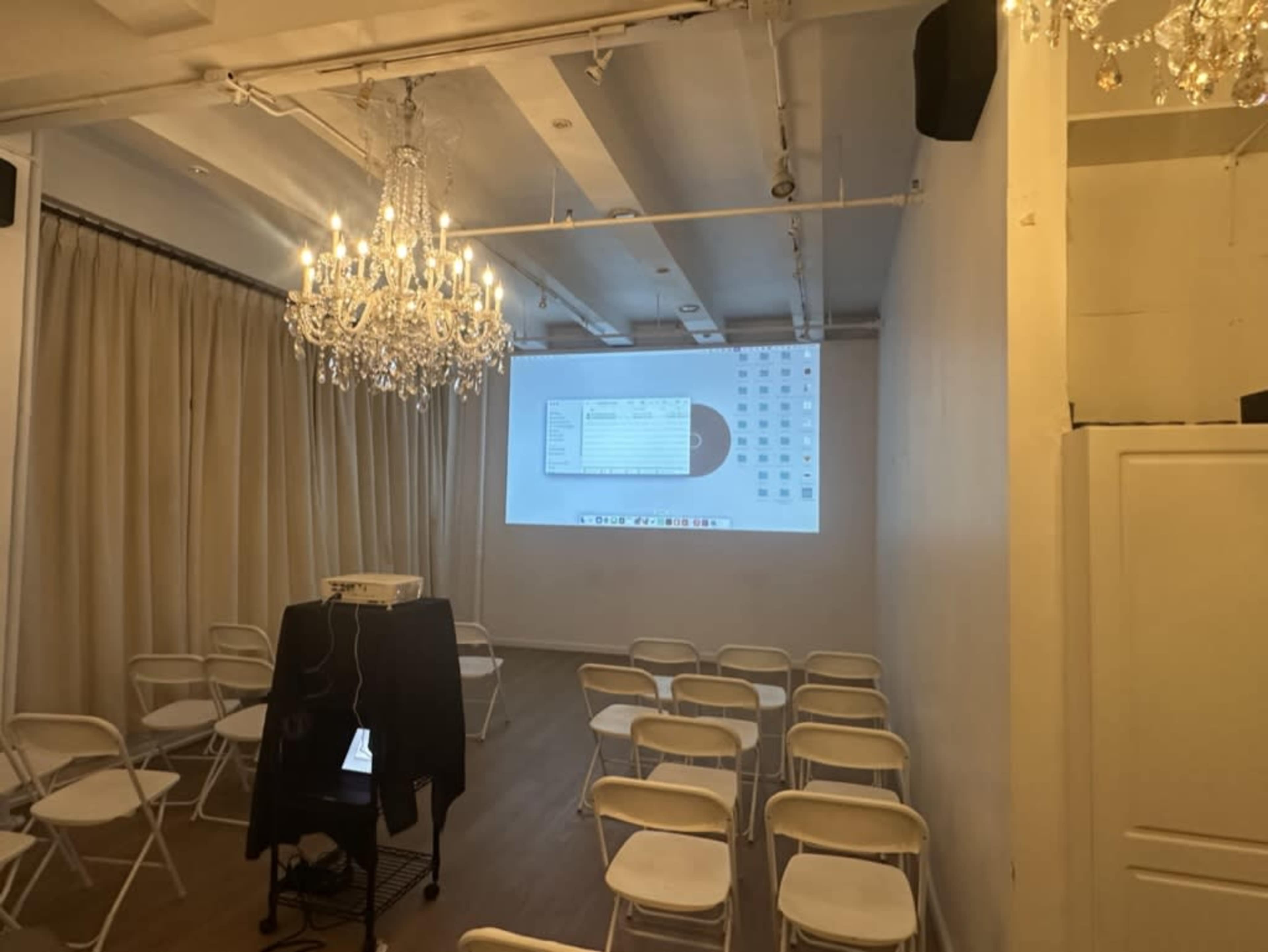 The image shows an indoor space set up for a presentation, featuring white folding chairs arranged in rows, a projector displaying a screen, and a chandelier hanging from the ceiling.