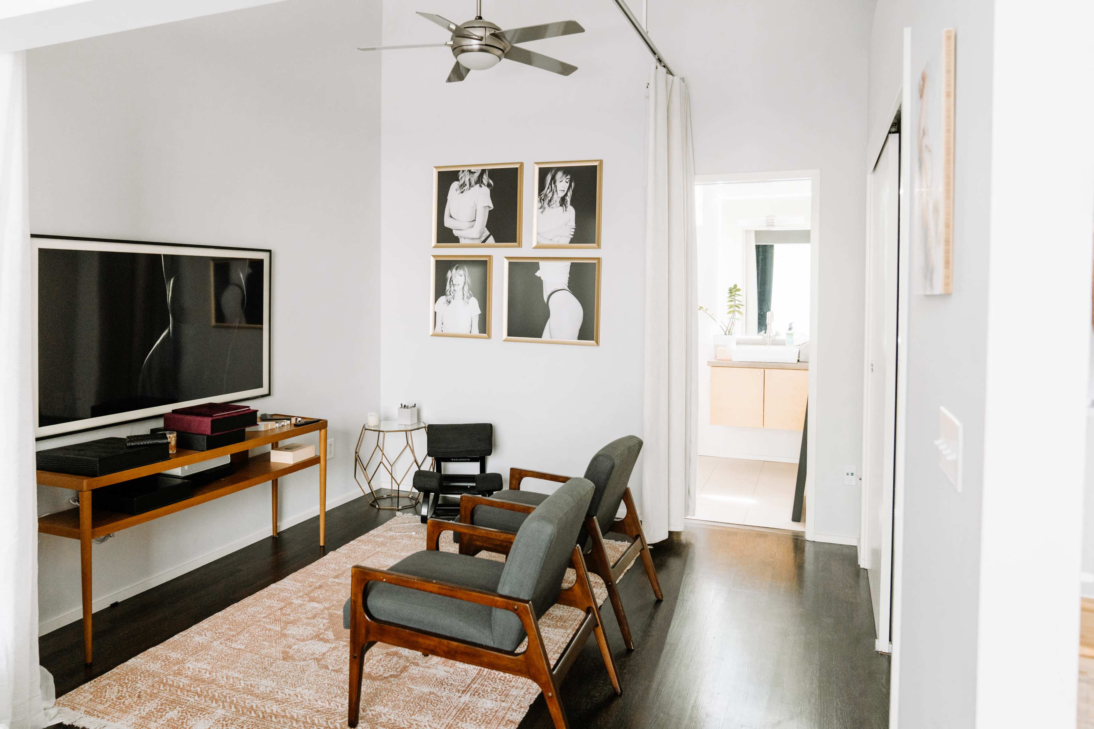 A modern living room features a television mounted on the wall, a wooden table with decorative items, three armchairs, and a hallway leading to a bathroom.