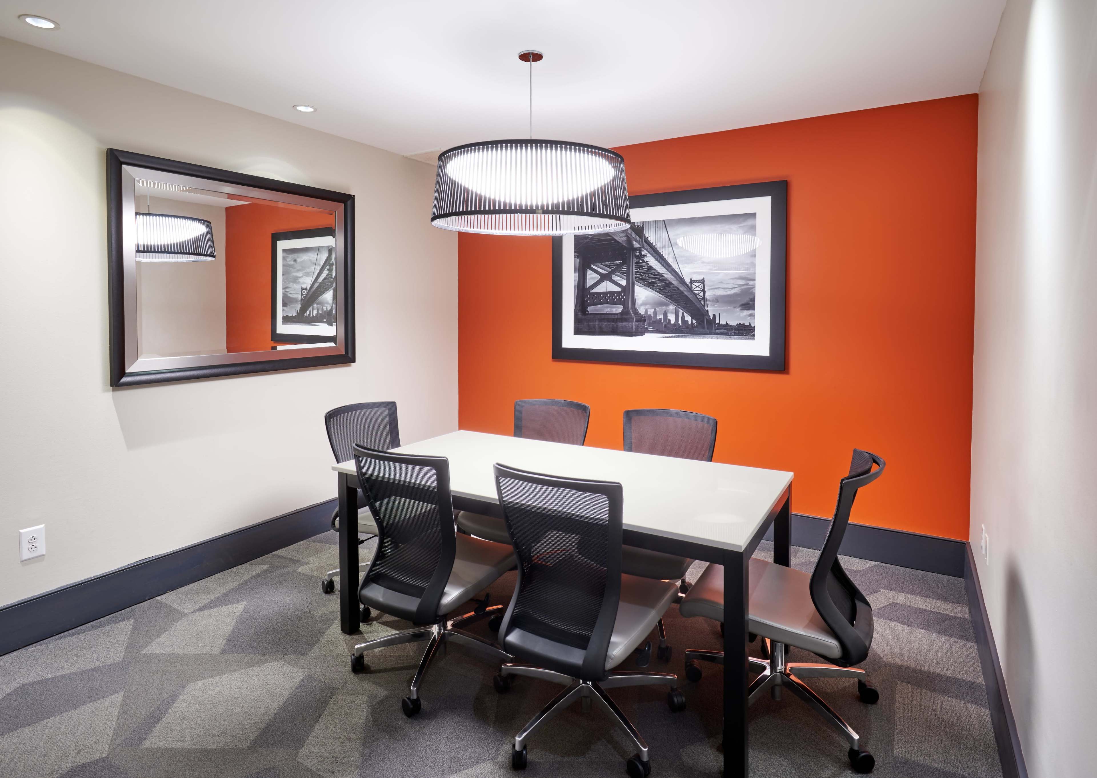 The image shows a modern conference room with a black table, six ergonomic chairs, an orange accent wall, and a large mirror reflecting the space.