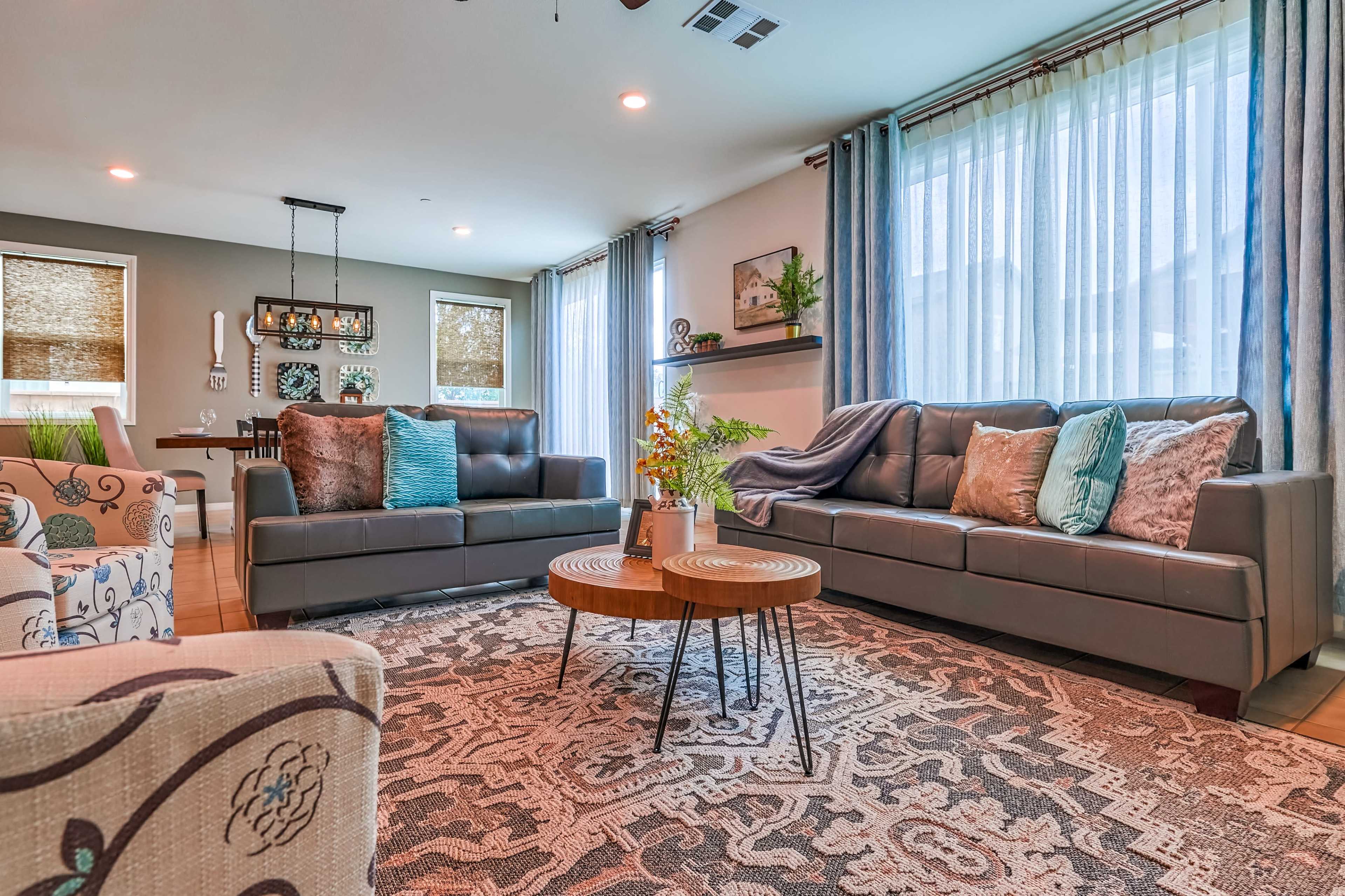 The living room features two gray sofas facing each other, with a round coffee table set on a patterned rug and large windows dressed in sheer curtains.