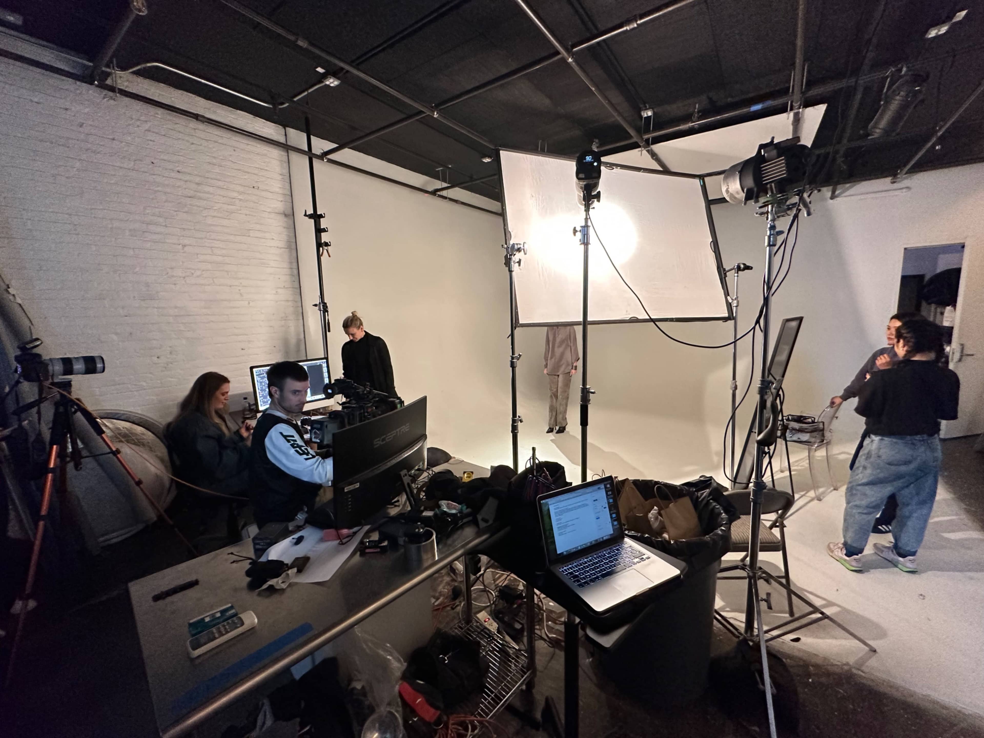 A photography studio is set up with several lights, a backdrop, and a team of individuals working at computer stations while one person stands in front of the backdrop.