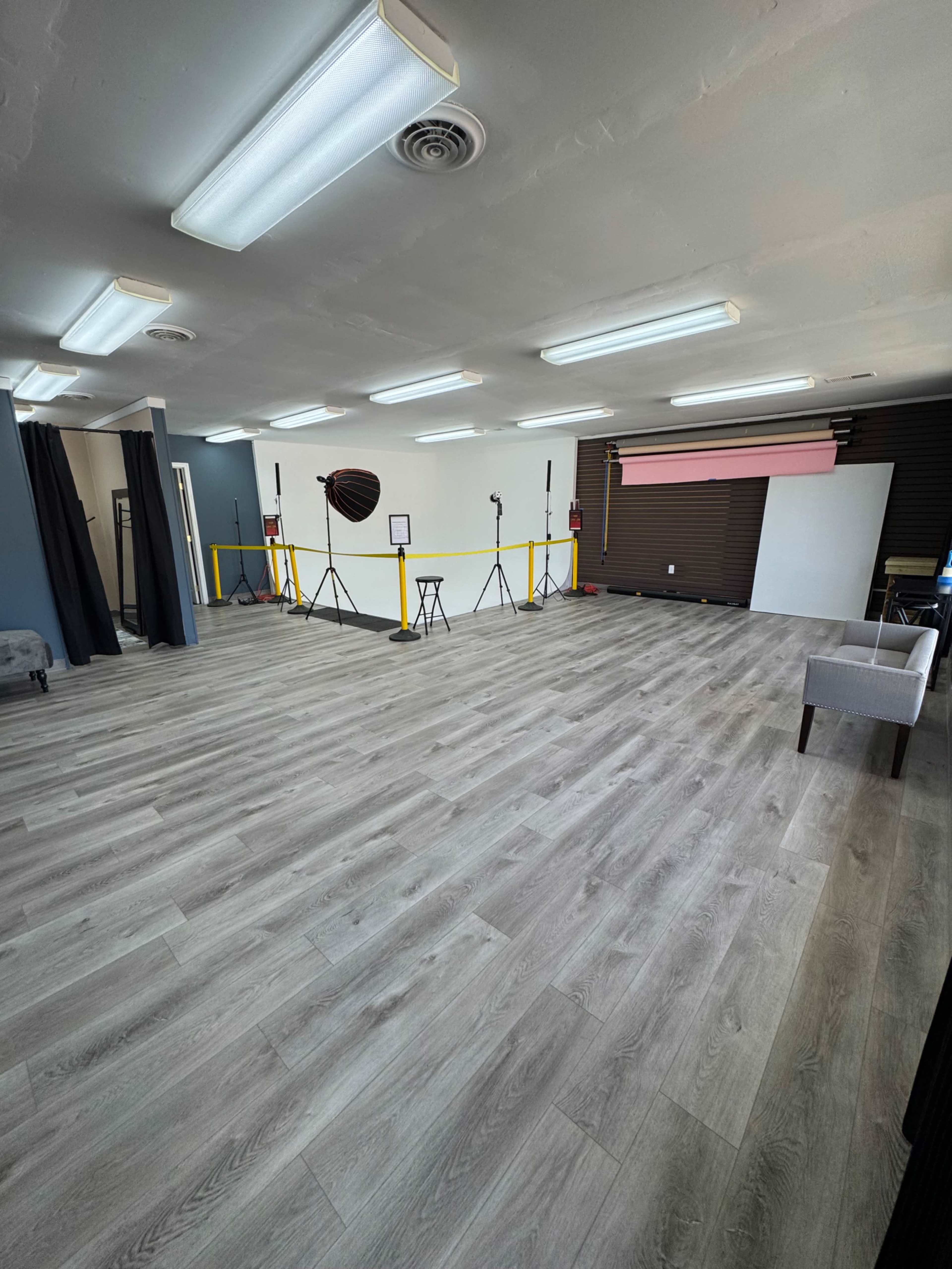 An empty studio space features wooden flooring, several lighting setups, and portable backdrops against a wall.
