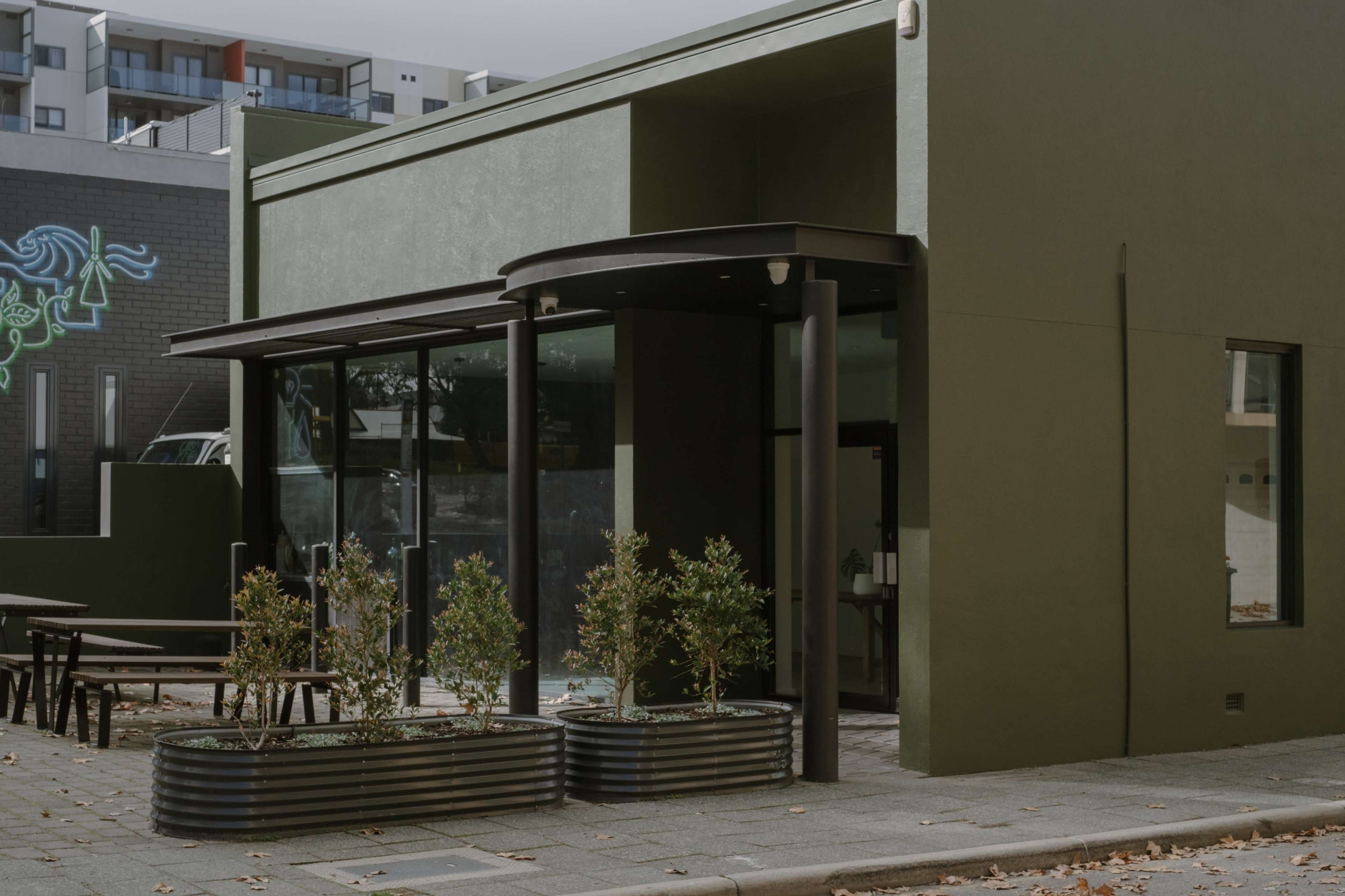 A modern green building with large glass windows and planters, located at the corner of a street.