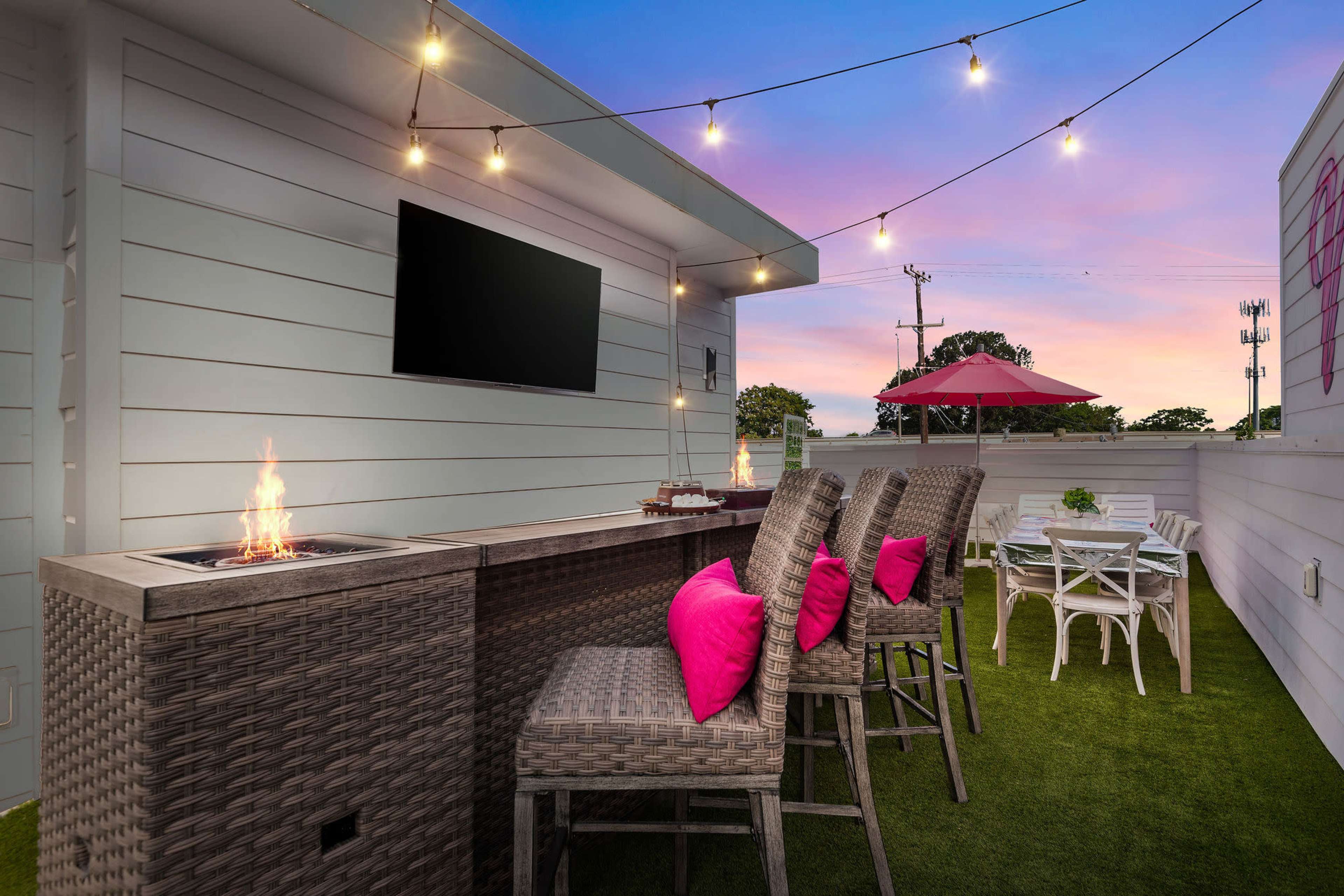 The image shows a rooftop patio with a fire pit, bar seating, and a dining area, accented by string lights and a pink sunset in the background.