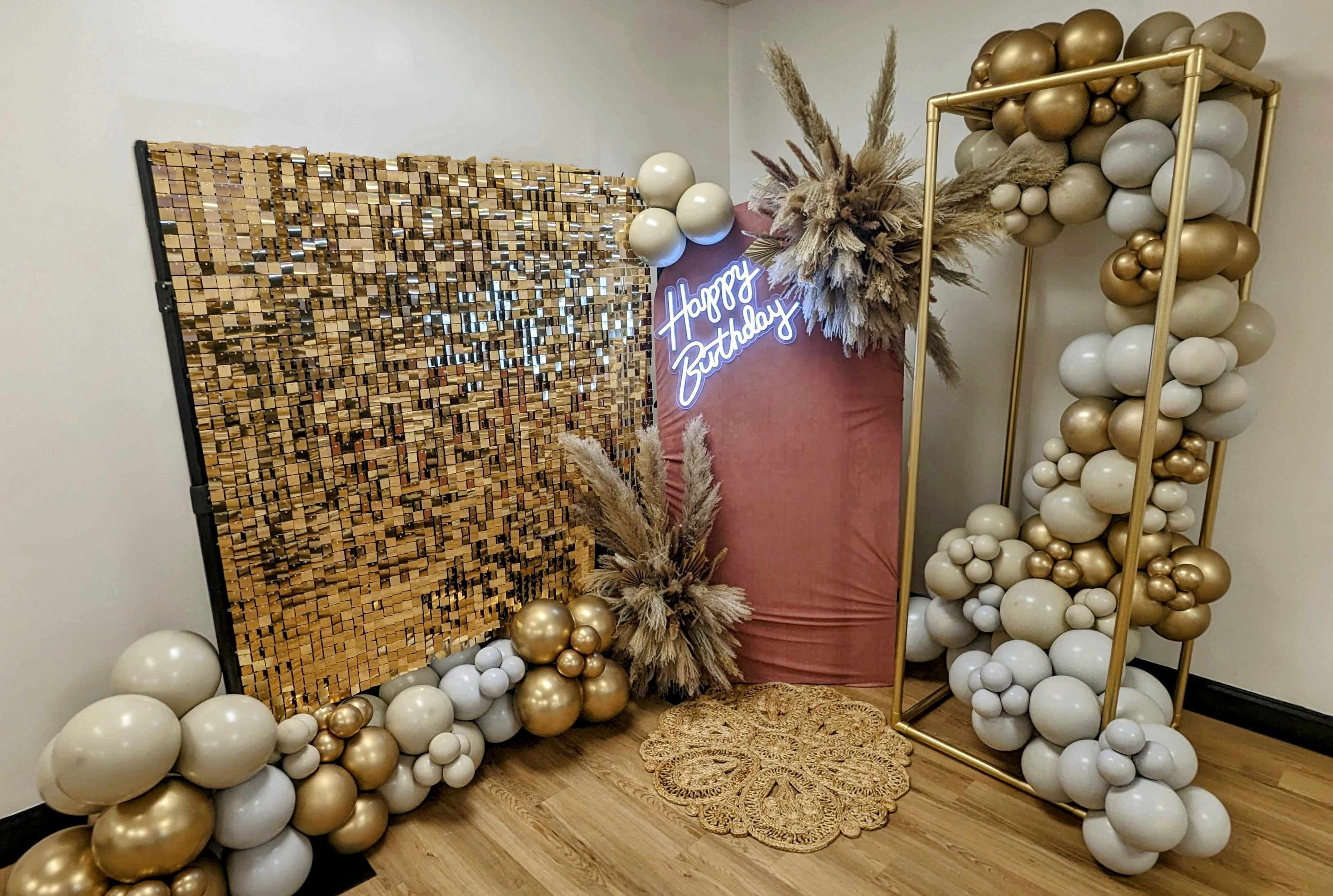 The image shows a birthday celebration setup featuring a gold sequin backdrop, a pink display with a neon "Happy Birthday" sign, a balloon arch, and decorative elements like pampas grass and a lace table piece.