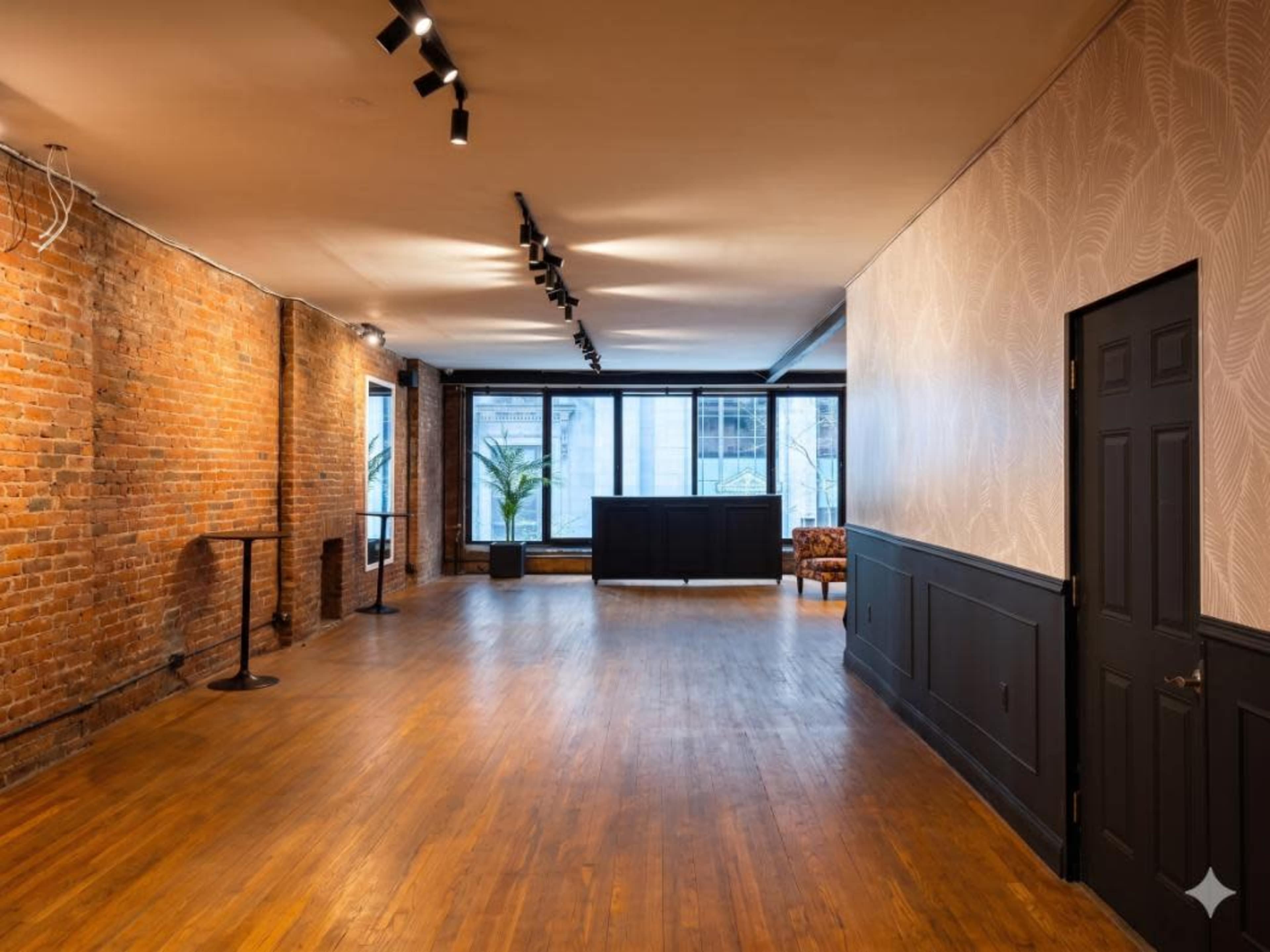 The image shows a spacious room with exposed brick walls, wooden flooring, and large windows, featuring a reception area and minimal furniture.