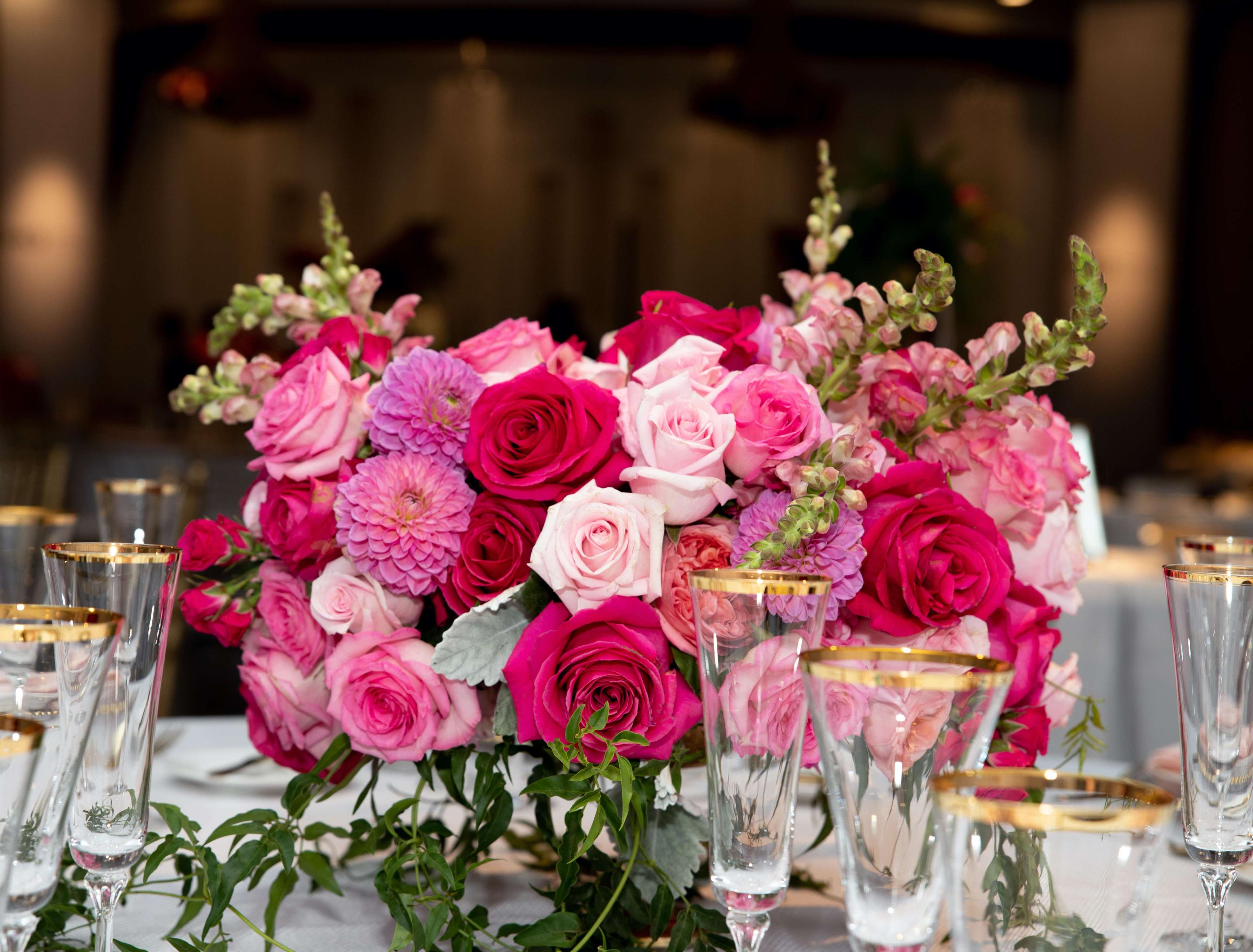 A vibrant floral arrangement featuring pink and red roses, zinnias, and snapdragons is displayed at a dining table surrounded by elegant glassware.