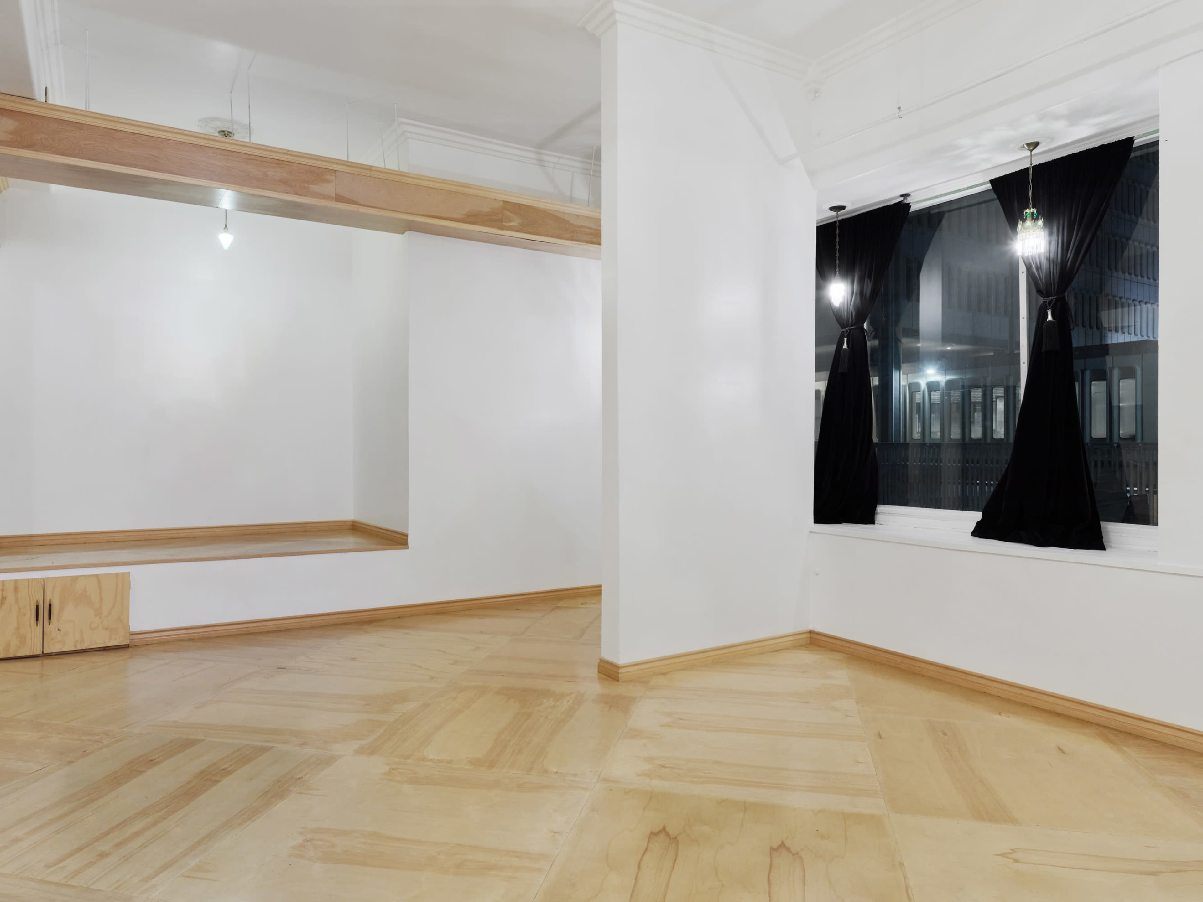 The image shows a spacious, minimalist room with wooden flooring, large windows draped in black curtains, and a simple wooden shelf along one wall.