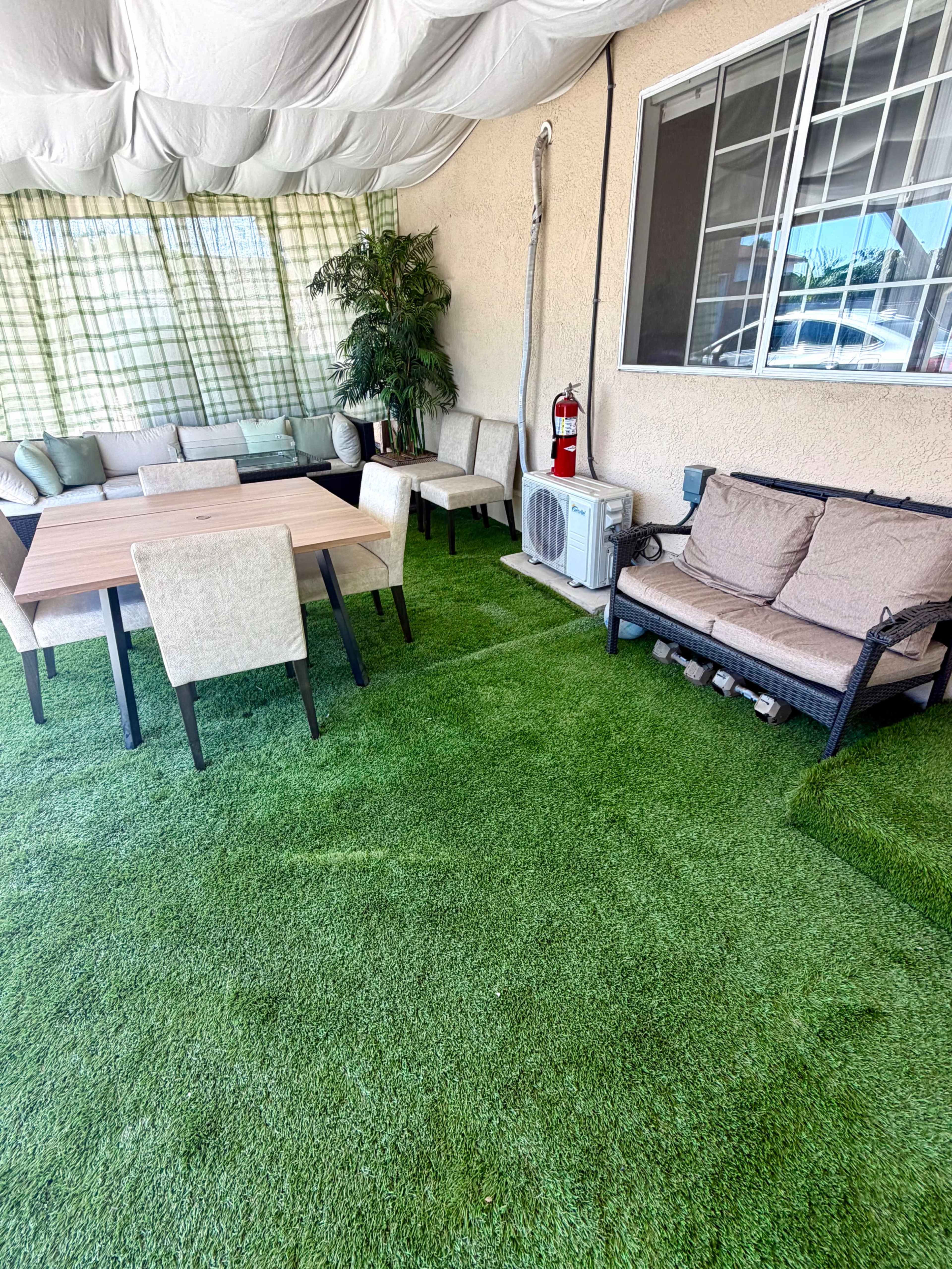 The image shows a covered outdoor seating area with a green artificial lawn, a wooden table surrounded by chairs, and a couple of couches.