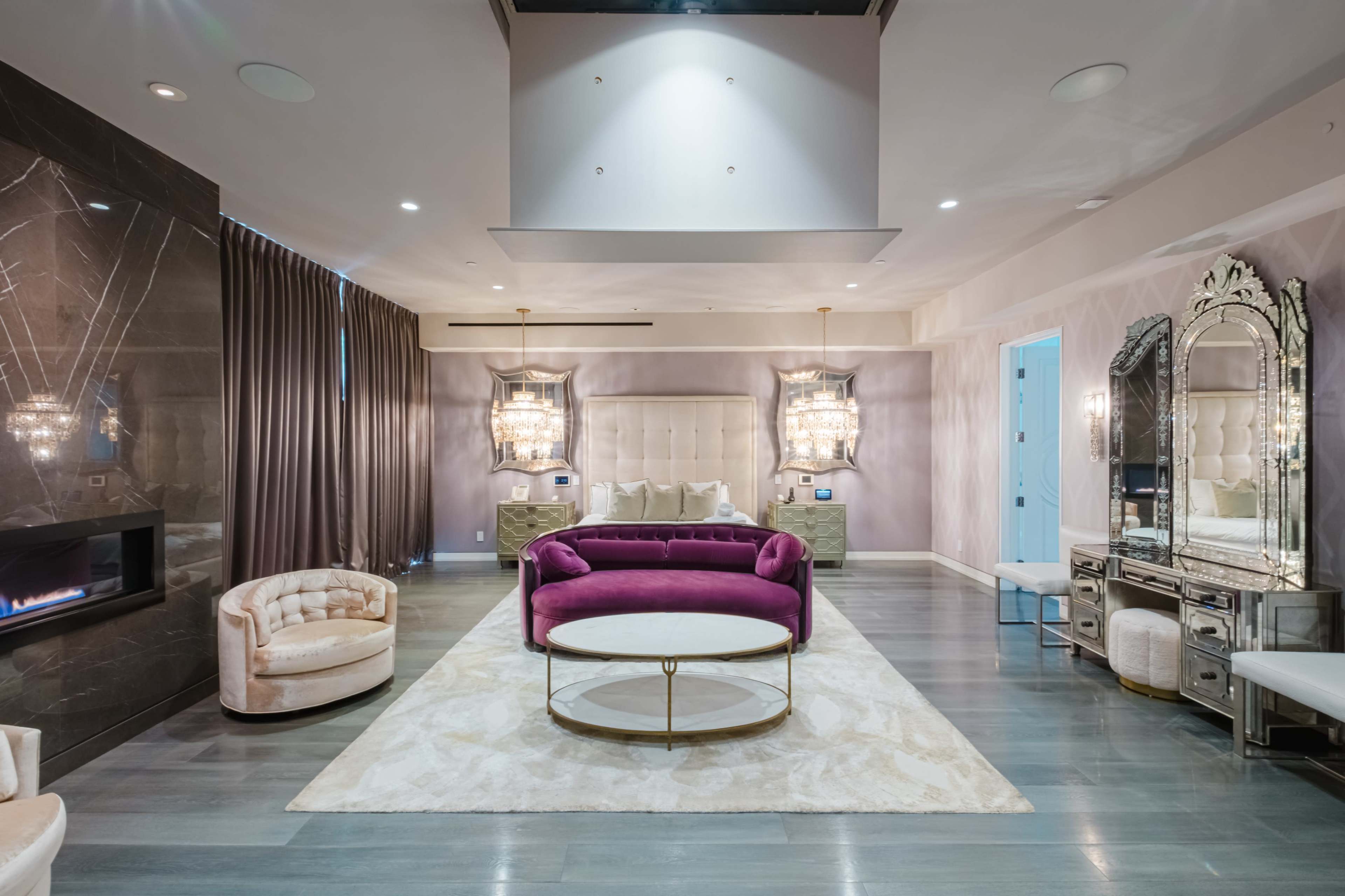 The scene features a modern living room with a purple velvet sofa, a round coffee table, and elegant decor elements, including a mirrored dresser and overhead chandeliers.
