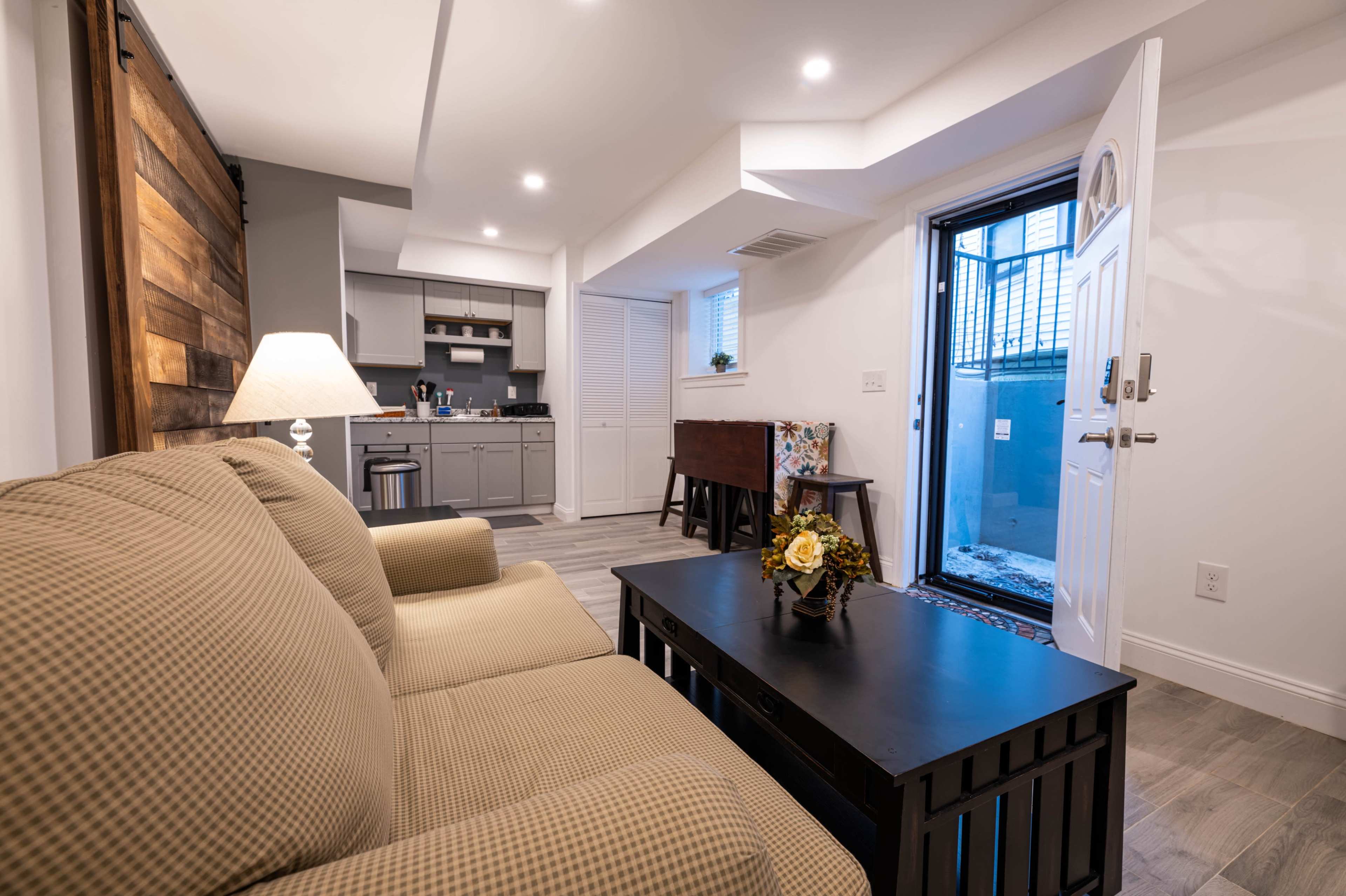 The image shows a cozy living space with a sofa, a coffee table, and a door that leads to an outdoor area, featuring a small kitchenette in the background.