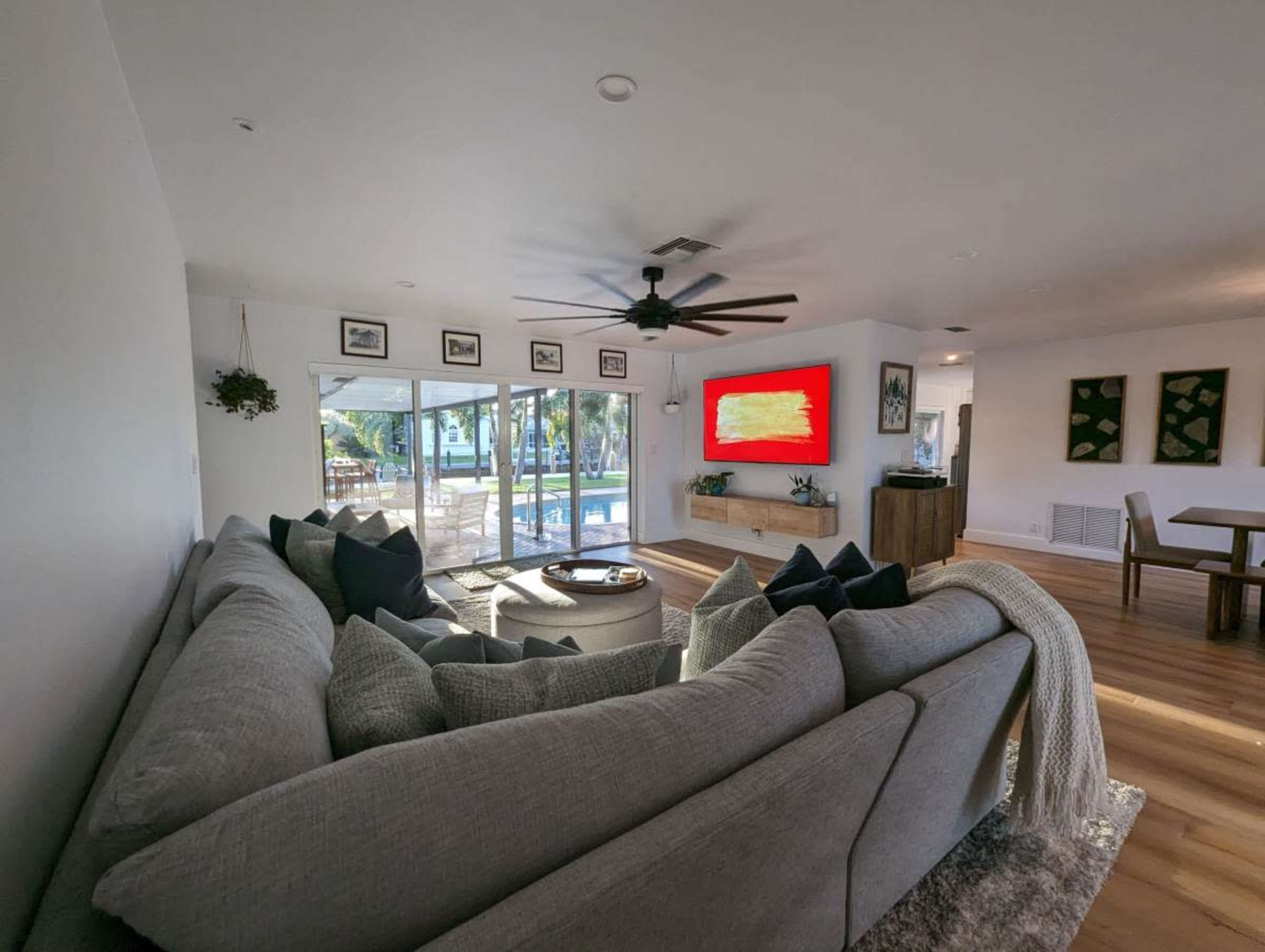 A modern living room features a large sectional sofa, a coffee table, and an abstract red artwork on the wall, with sliding glass doors opening to a pool area outside.