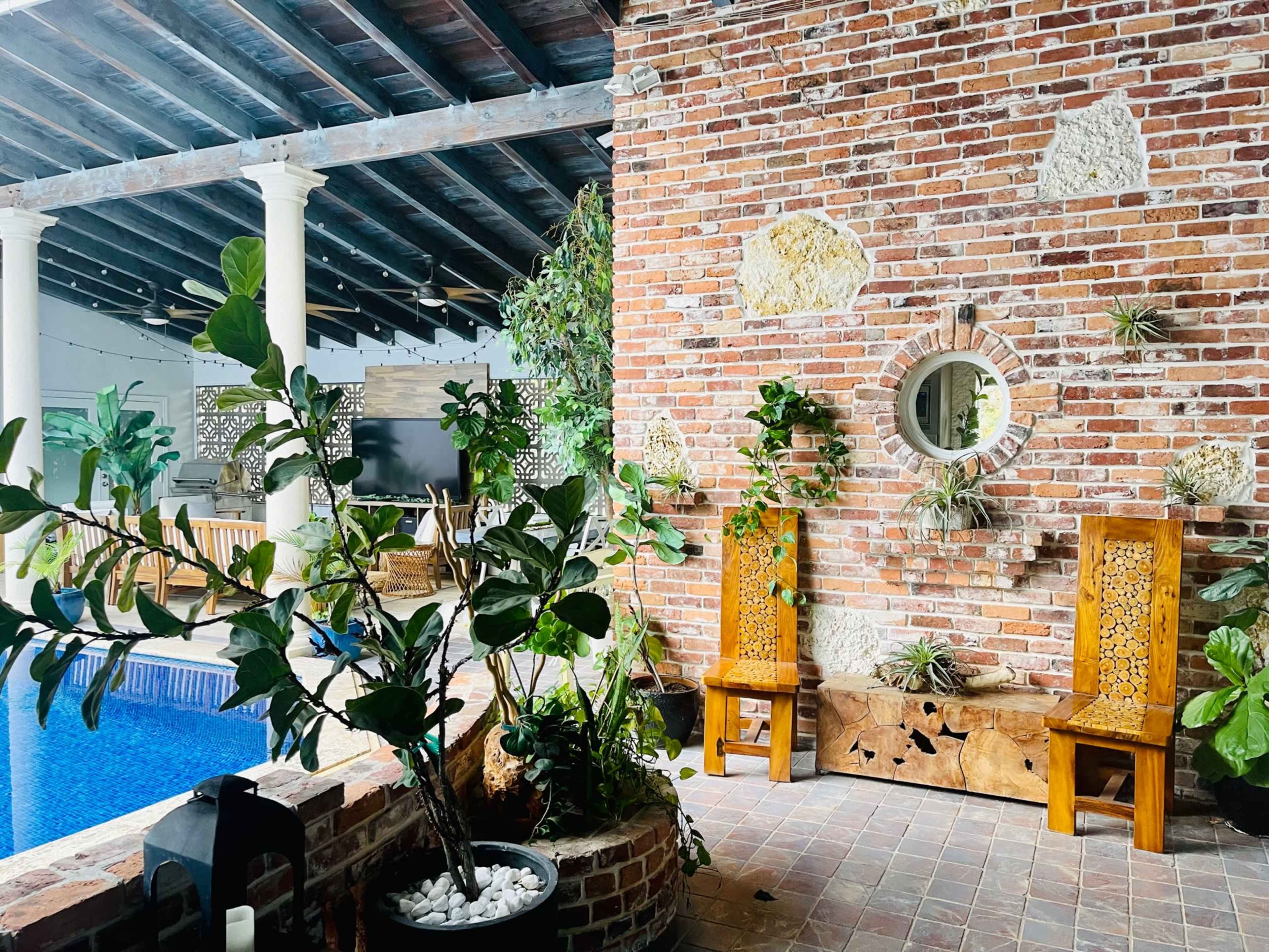 An outdoor area with a swimming pool, a brick wall featuring decorative stone patterns, and wooden benches surrounded by indoor plants.