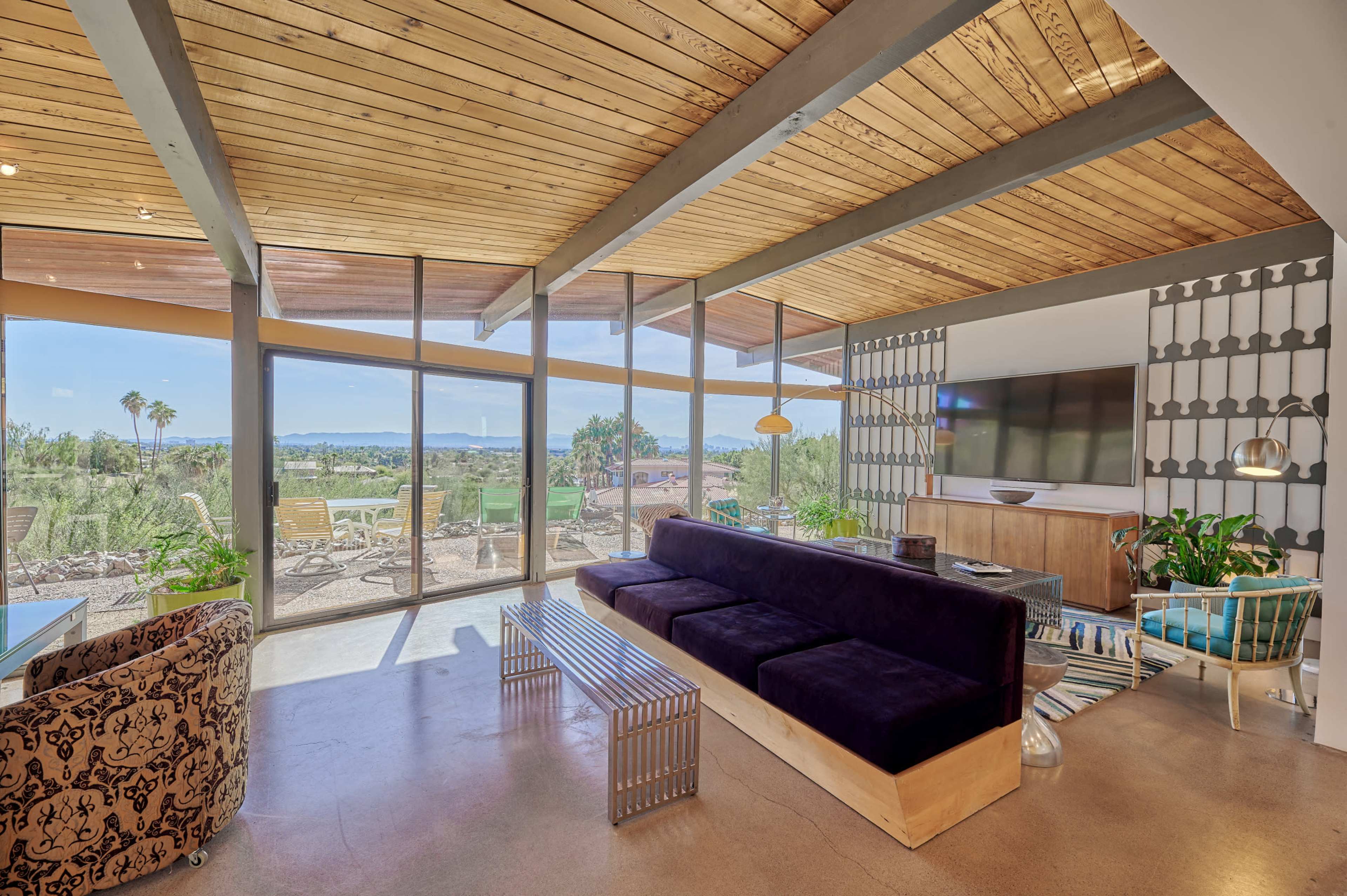 A modern living room features a large window with a view of the landscape, a dark sofa, a wooden bench, and a television on a wall unit.