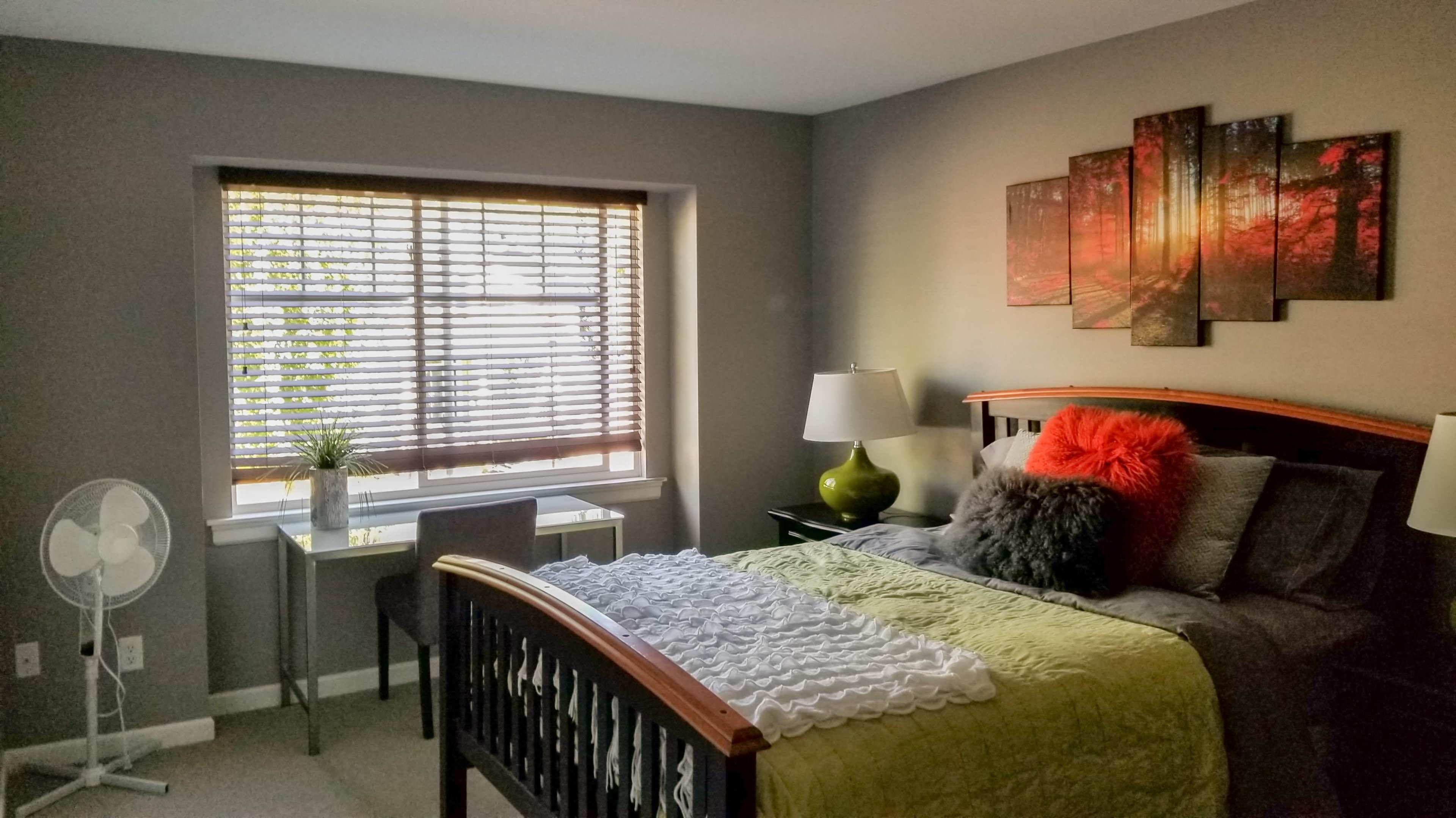 A well-furnished bedroom features a bed with decorative pillows, a window with blinds, a small desk and chair, and a fan.