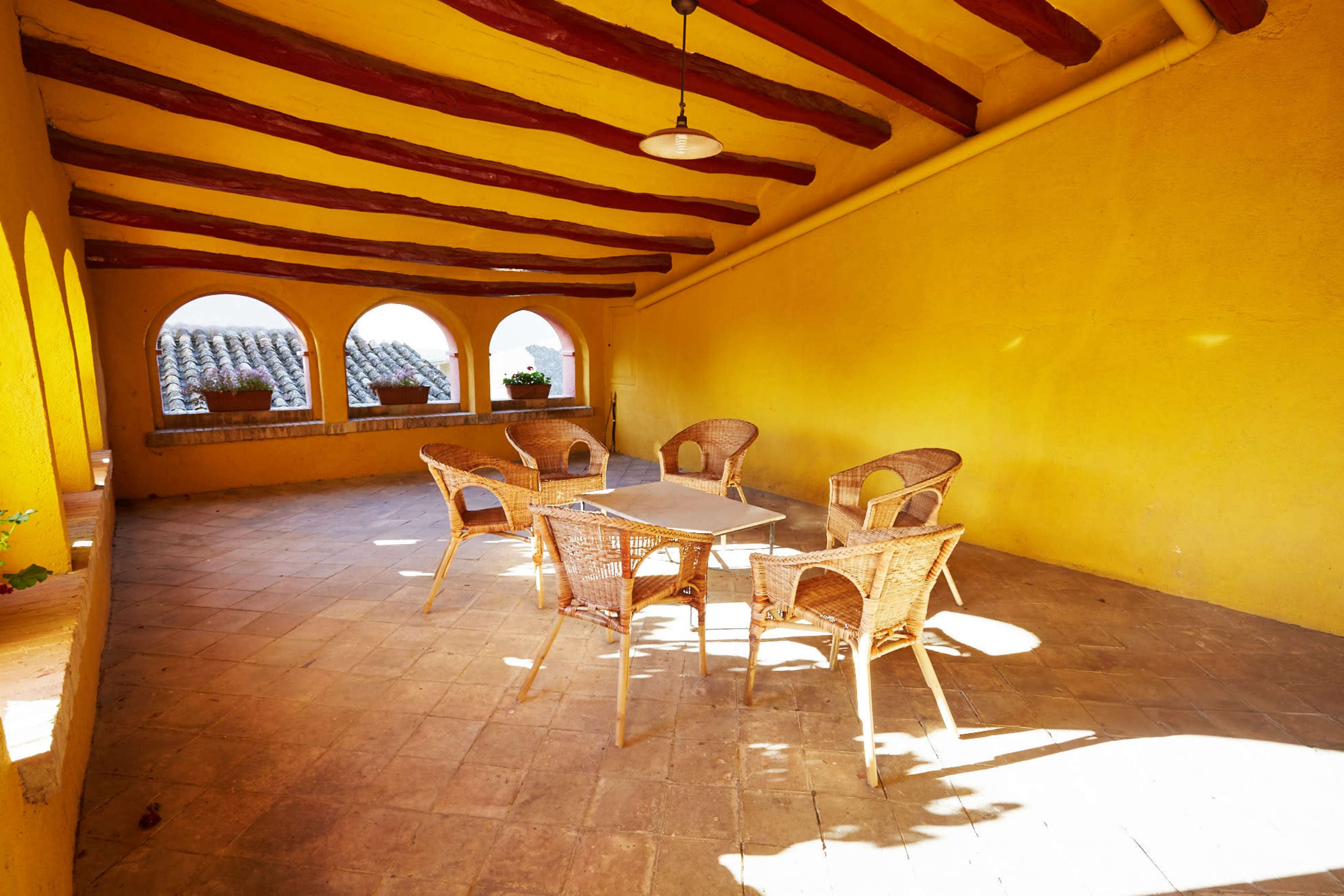 A sunlit patio with a table and four wicker chairs, surrounded by archways and potted plants.
