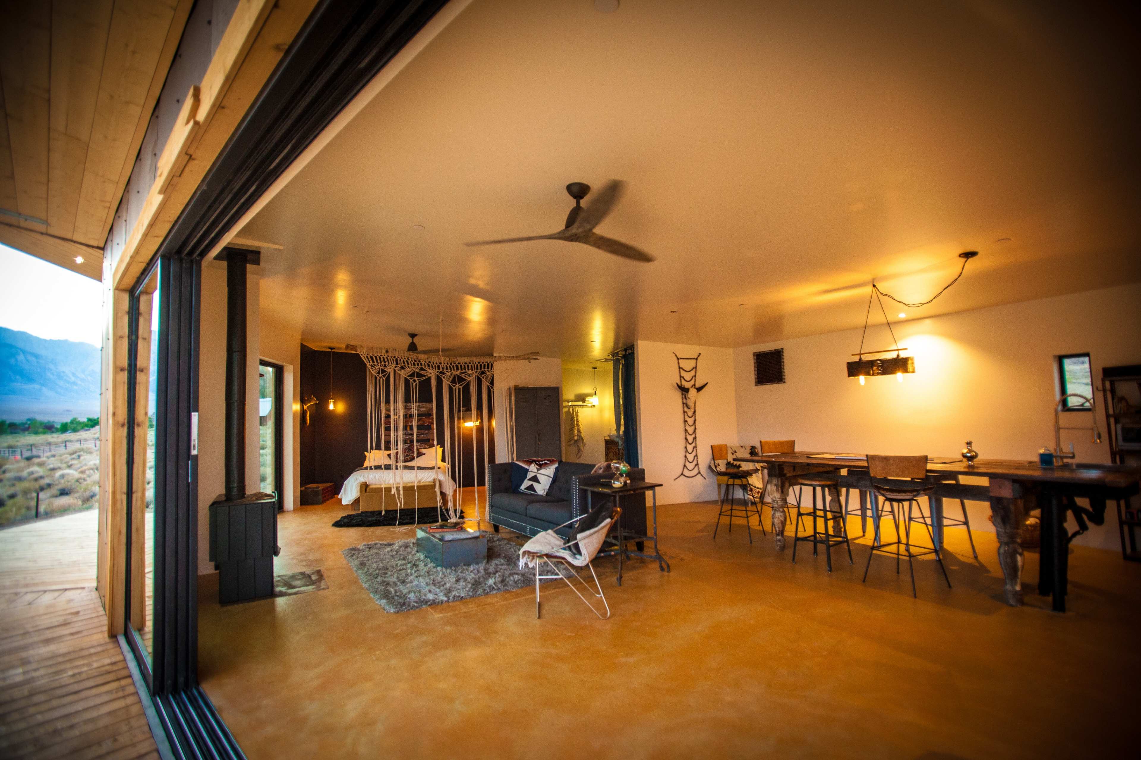 The image shows an open-concept living space featuring a dining area, a seating area, and a bedroom with a canopy bed, illuminated by pendant lighting and large windows.