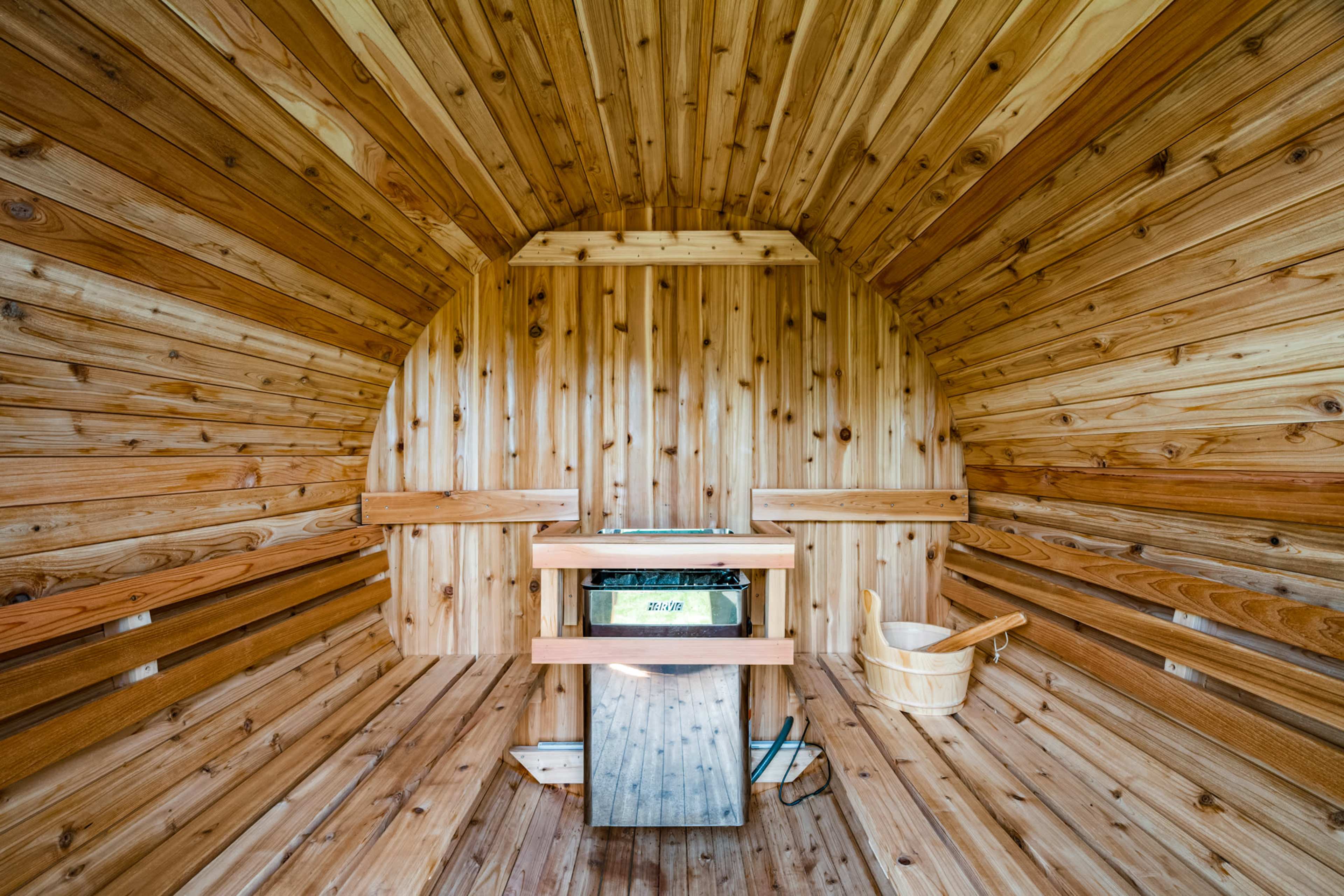 The interior of a wooden sauna features smooth, rounded cedar walls, a central bench, a stove, and a wooden bucket.