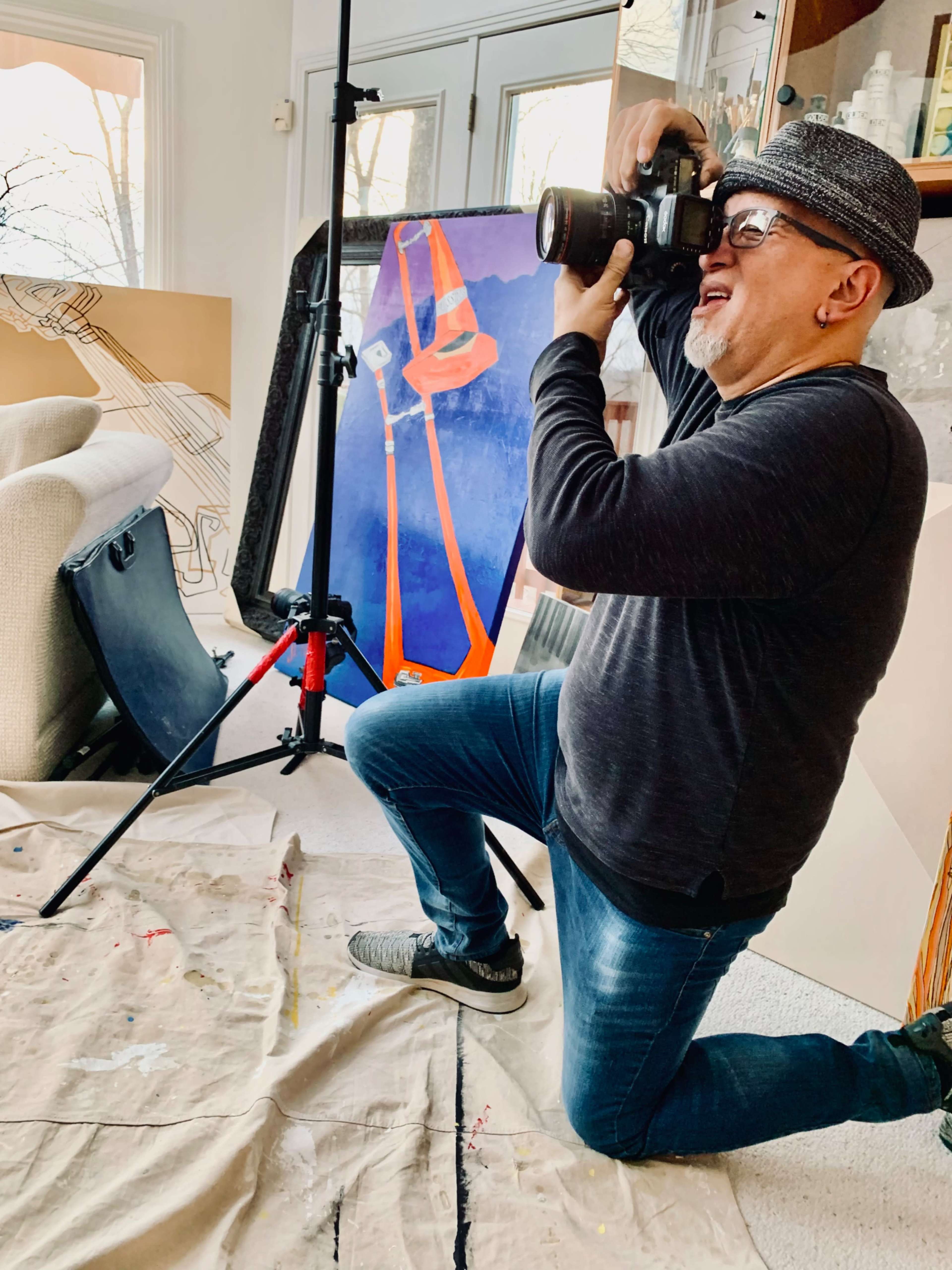 A photographer kneels on a cloth while aiming a camera at a colorful backdrop in a bright, cluttered studio.