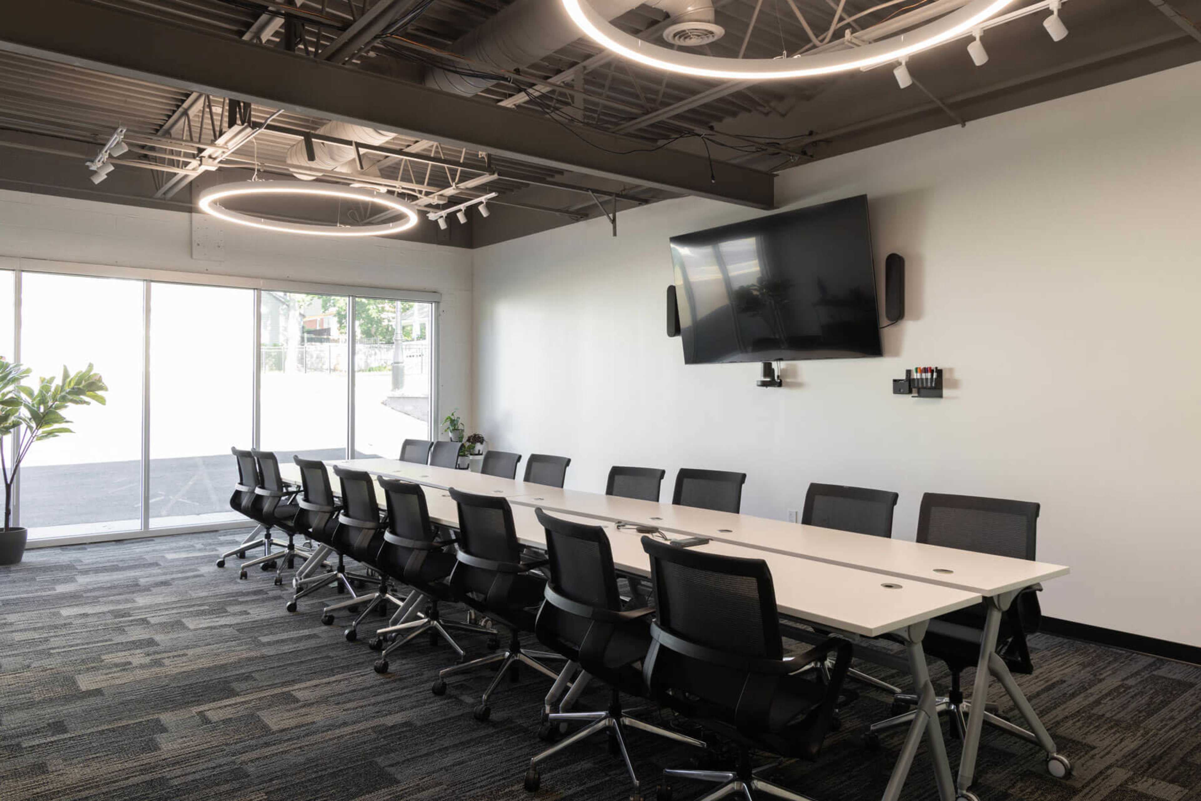 A modern conference room features a long table surrounded by black chairs, with a large flat-screen TV mounted on the wall and circular light fixtures overhead.