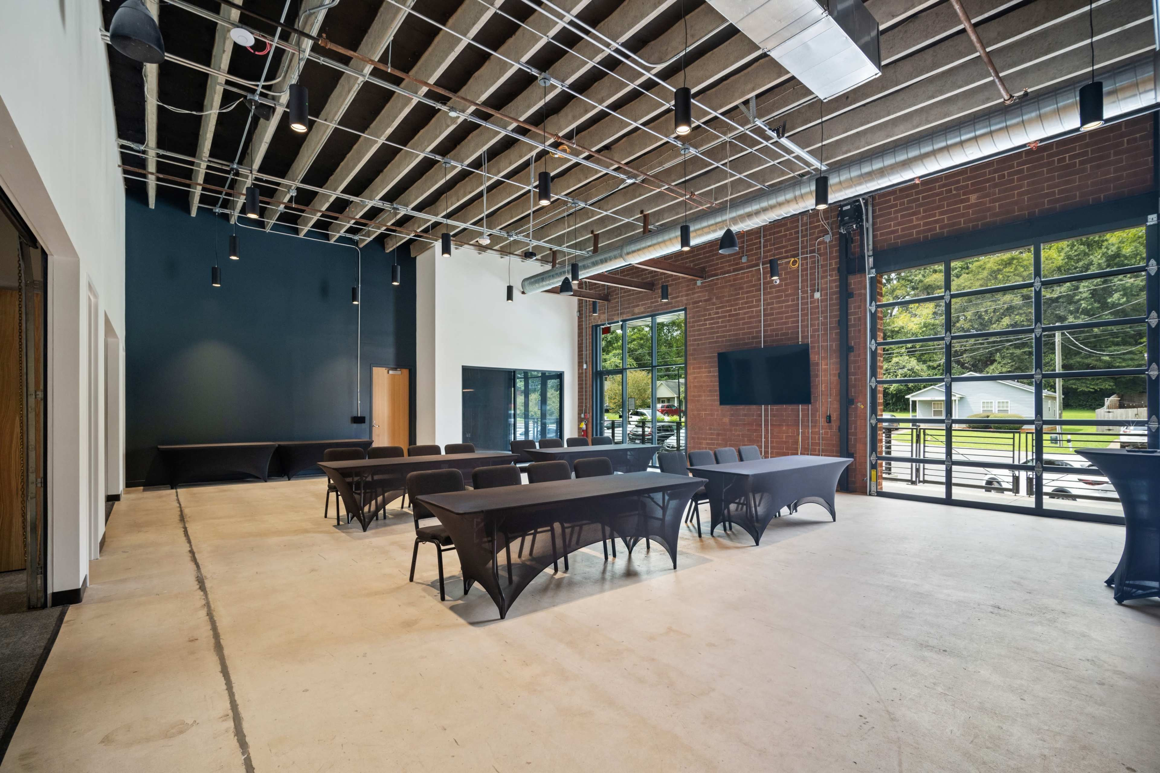 The image shows a spacious interior of a modern event room with large windows, a concrete floor, and several tables arranged for a meeting.