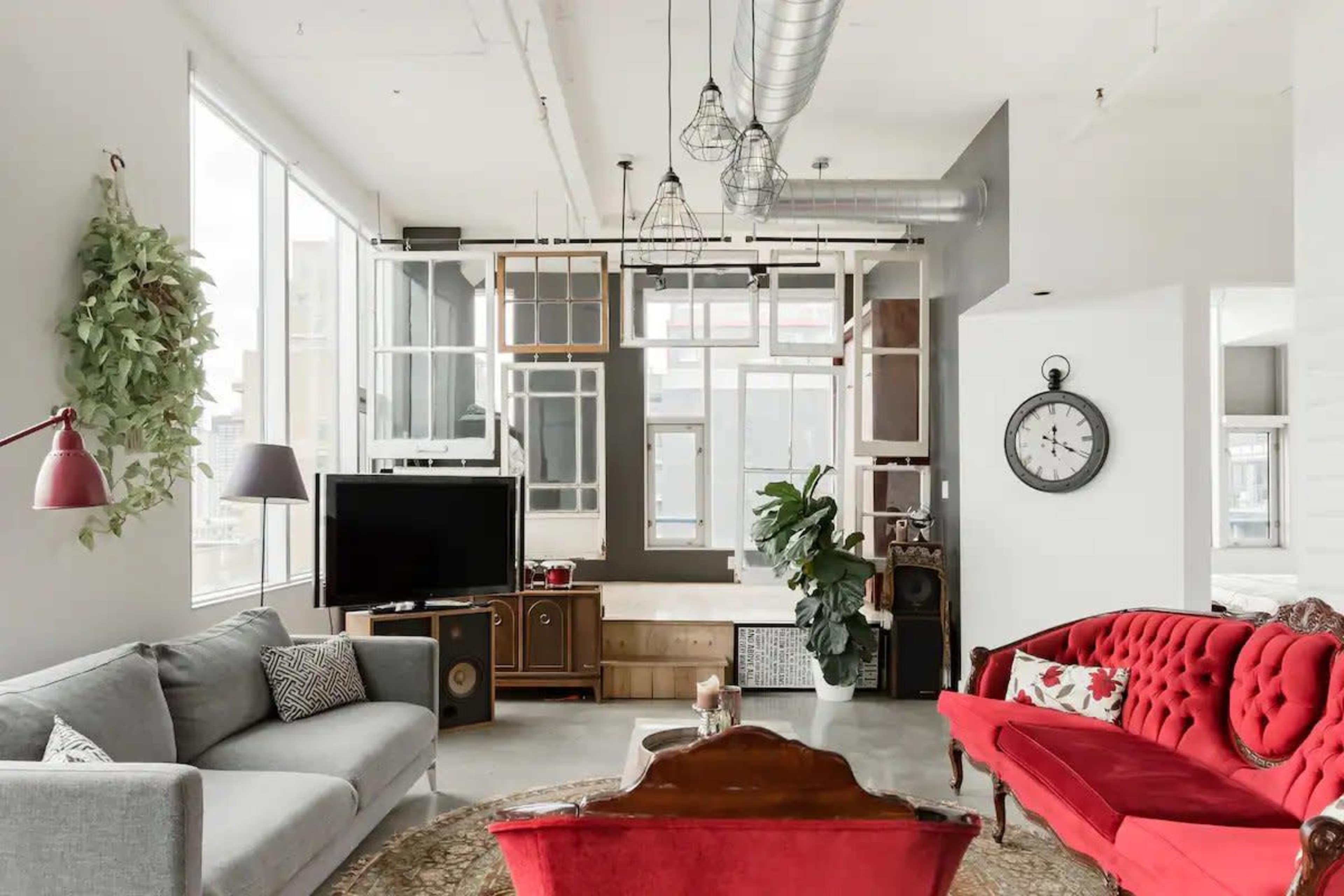 A modern living room features a gray sofa, a red vintage-style couch, a large television, and a mix of wall decorations including a clock and plants, with ample natural light from large windows.