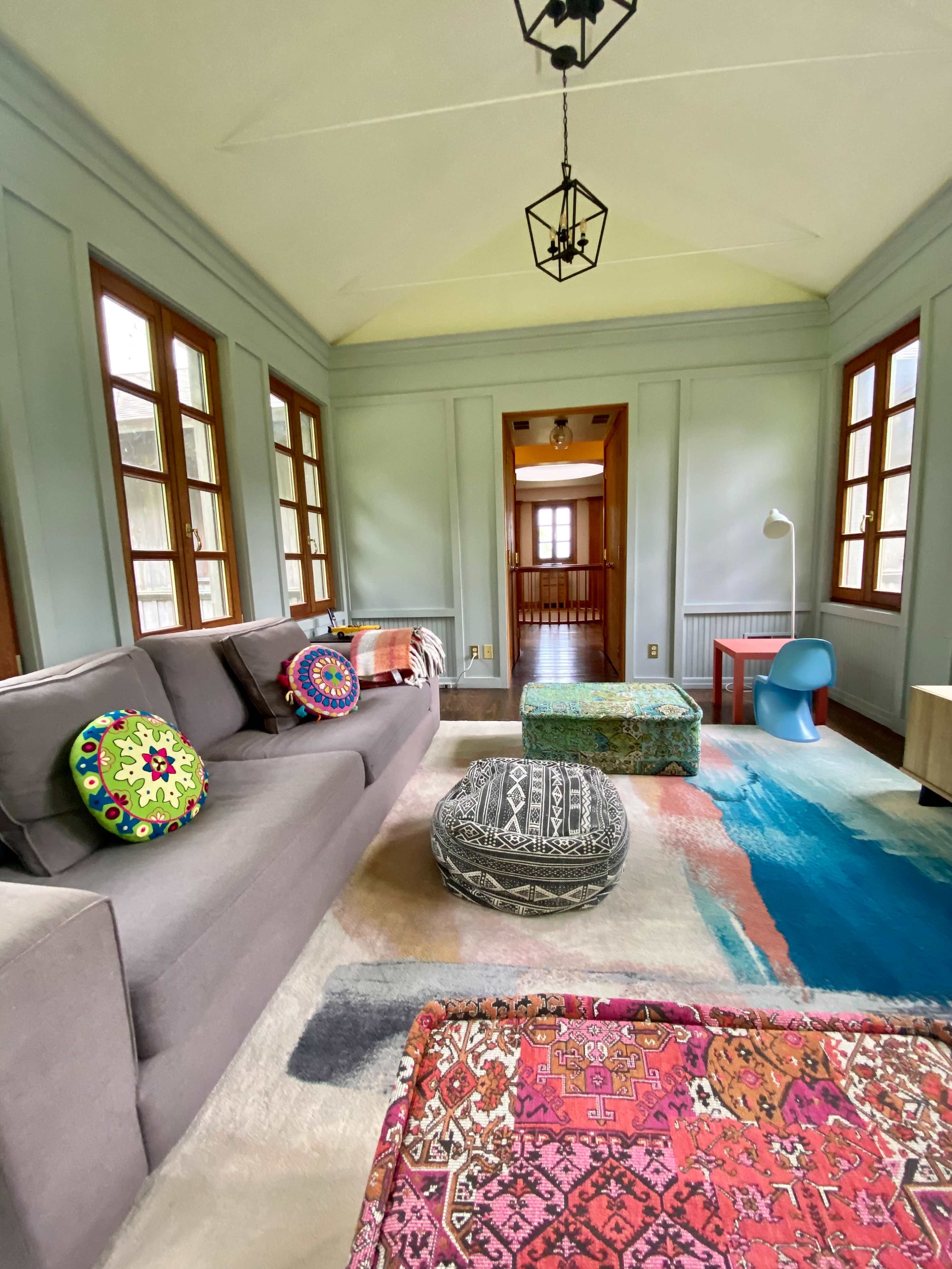 The room features a gray sofa with decorative cushions, a colorful rug, and large windows that let in natural light.