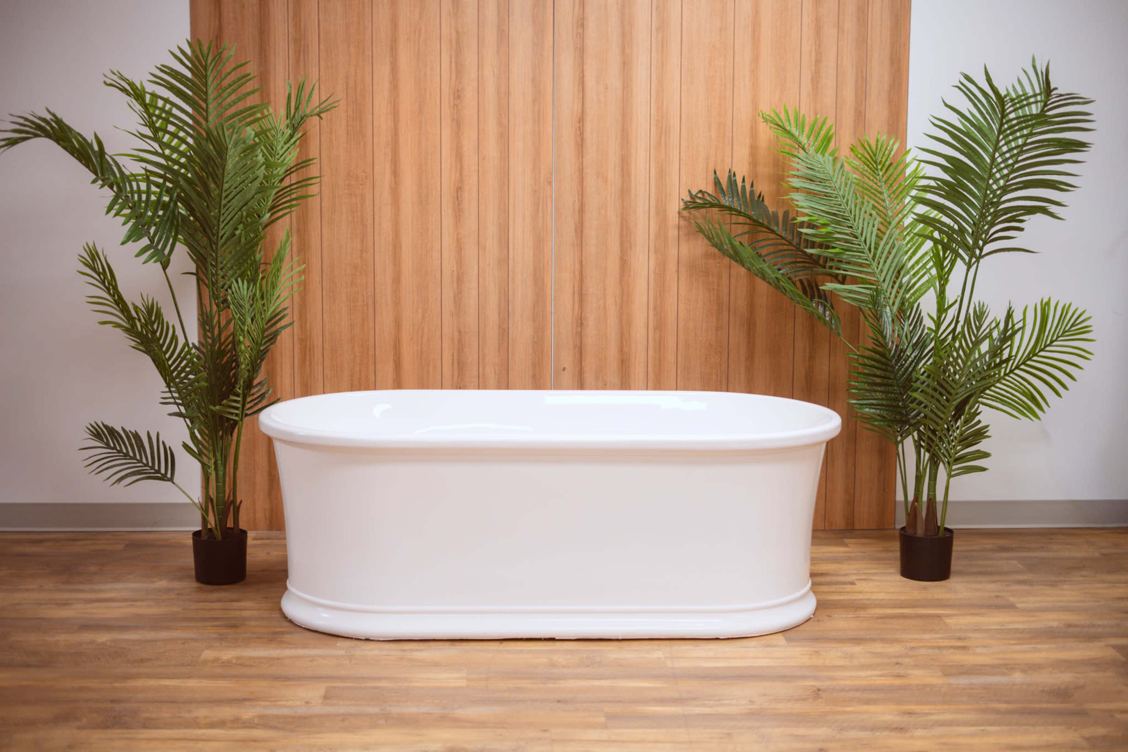 A white freestanding bathtub is positioned against a wooden backdrop, flanked by two large potted palm plants.