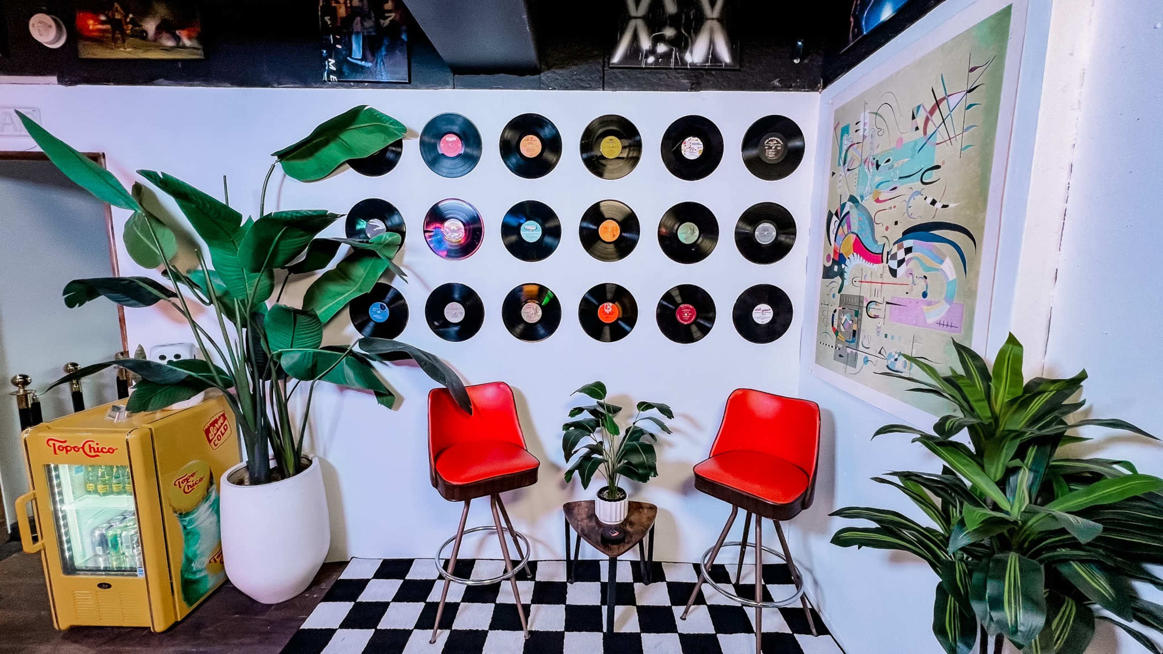The image shows a cozy corner of a room featuring a wall adorned with vinyl records, two red chairs, and potted plants, set against a checkered floor.