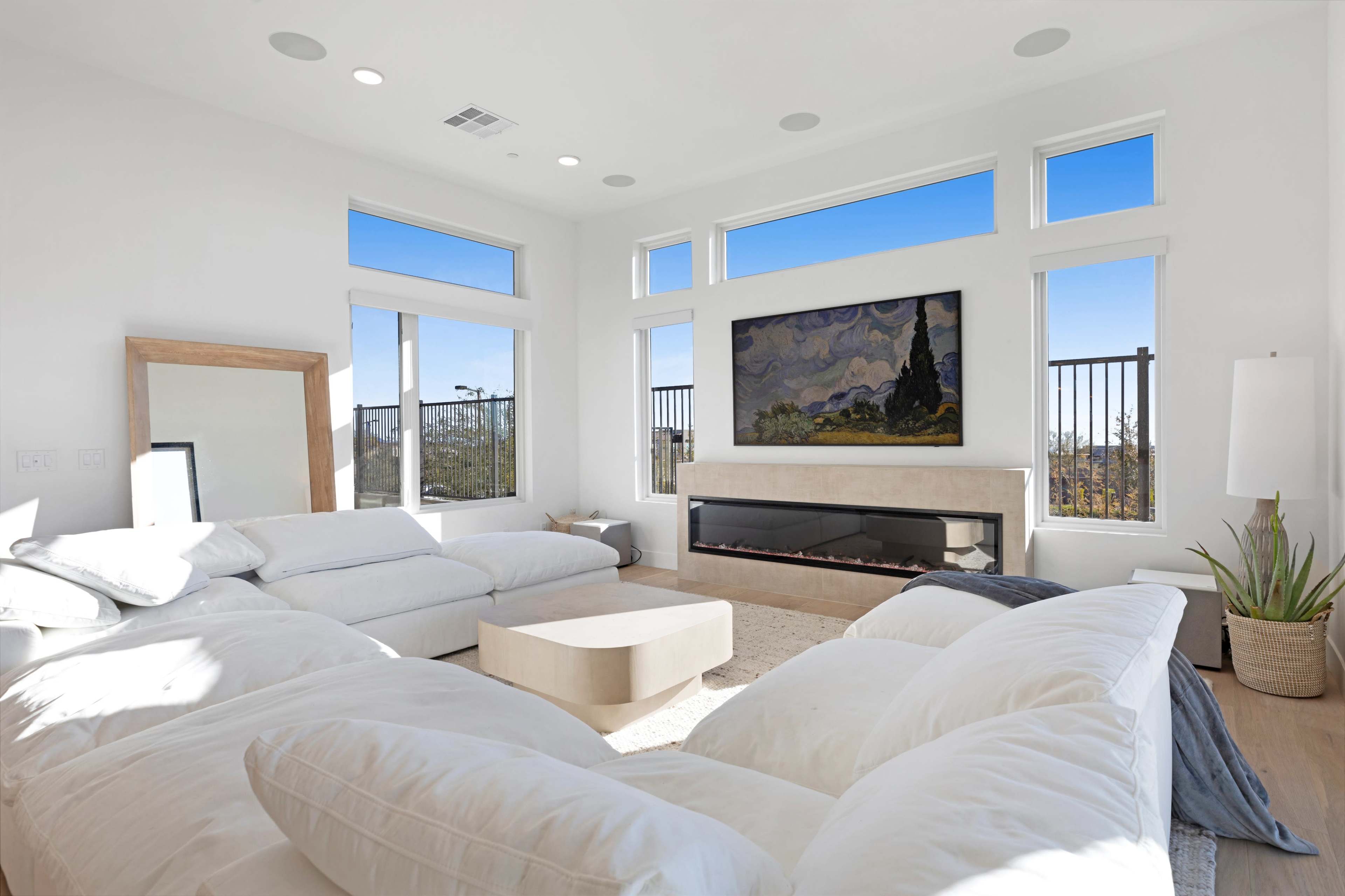 A modern living room features a large white sectional sofa, a minimal coffee table, and a wall-mounted artwork above a sleek fireplace, with ample natural light coming through large windows.