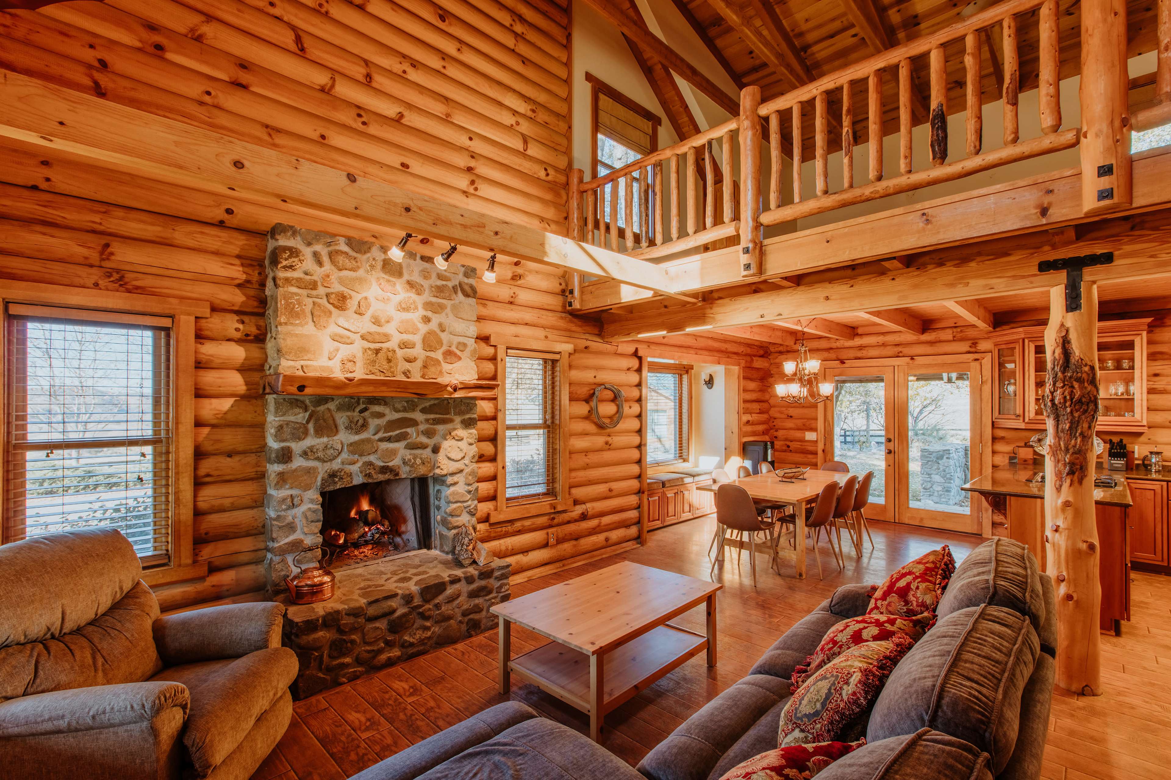 The interior of a rustic log cabin features a stone fireplace, wooden furniture, and a spacious open layout connecting the living area to a dining space.