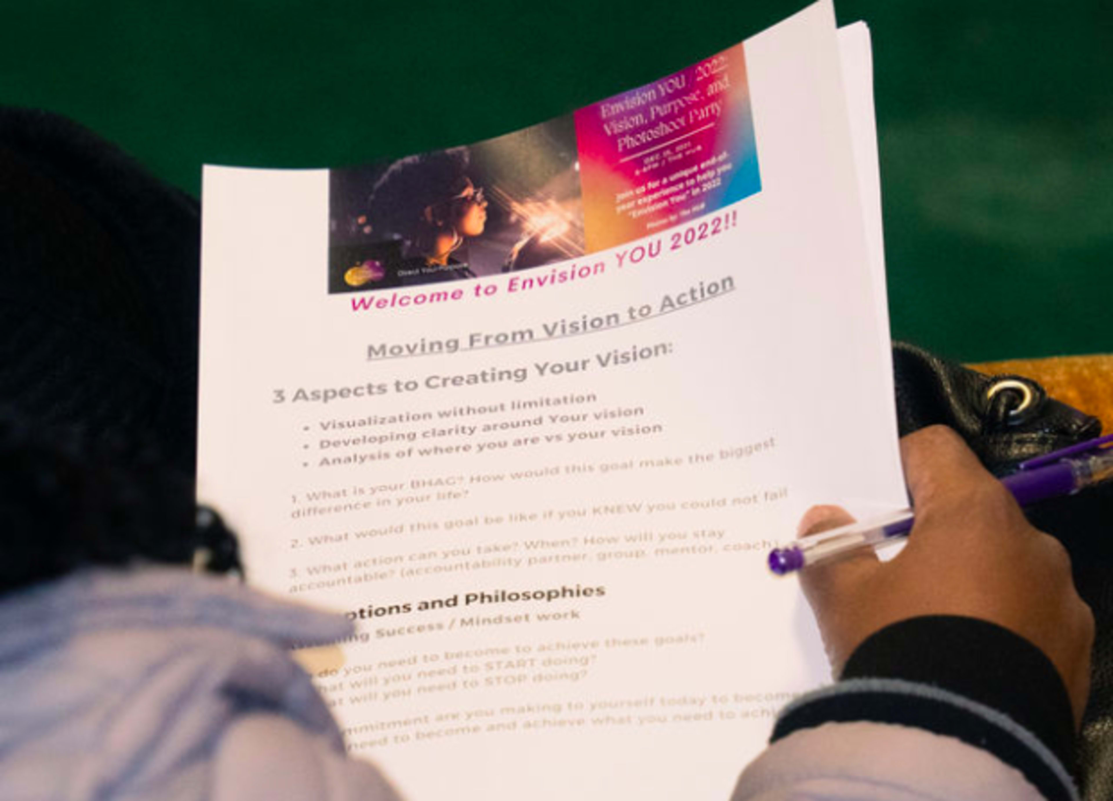 A person holds a printed handout titled "Welcome to Envision You 2022!" that outlines steps for creating a vision, including visualization, clarity, and goal setting.