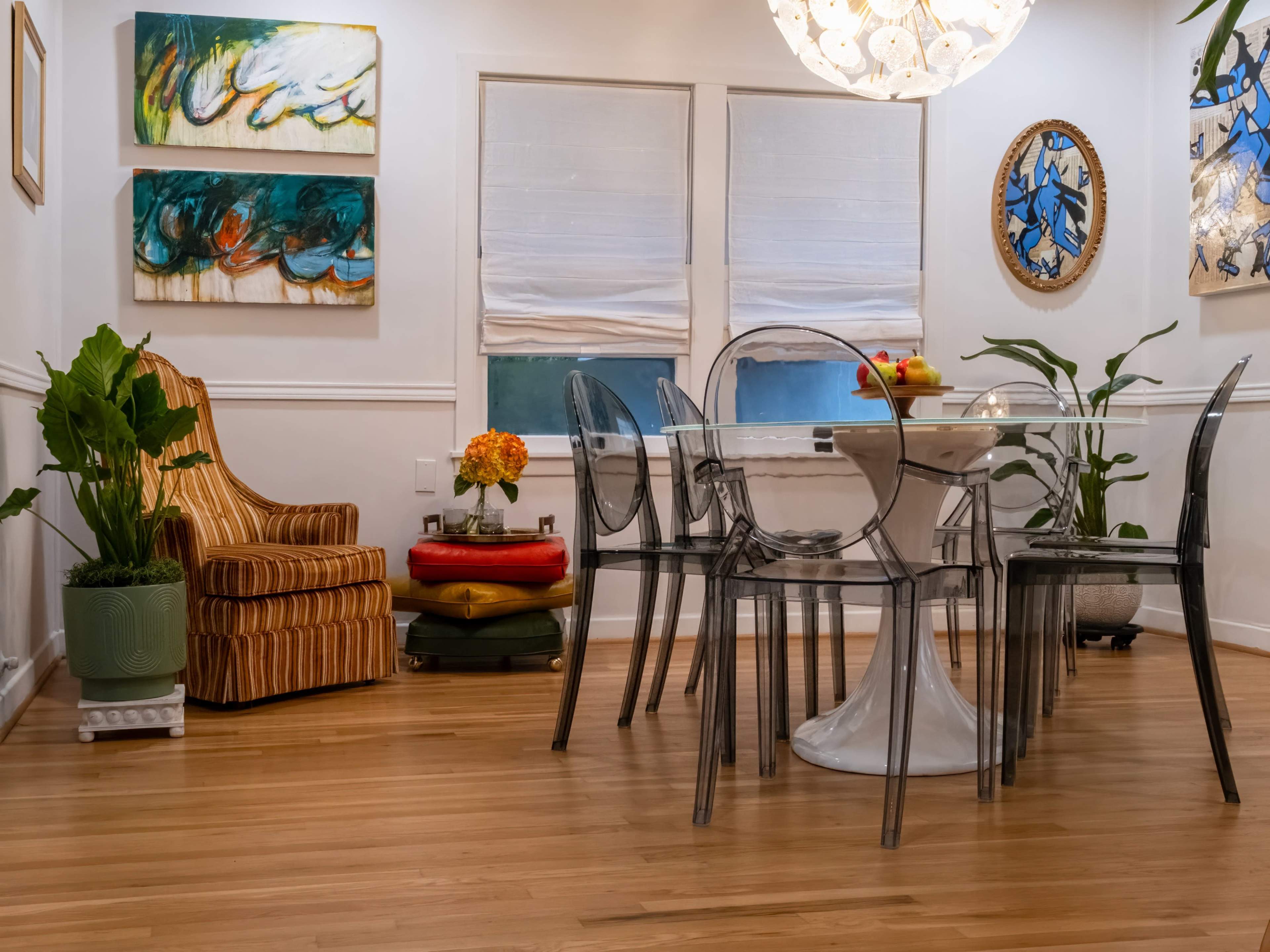 A dining area features a clear glass table surrounded by transparent chairs, a striped armchair, and vibrant artwork on the walls.